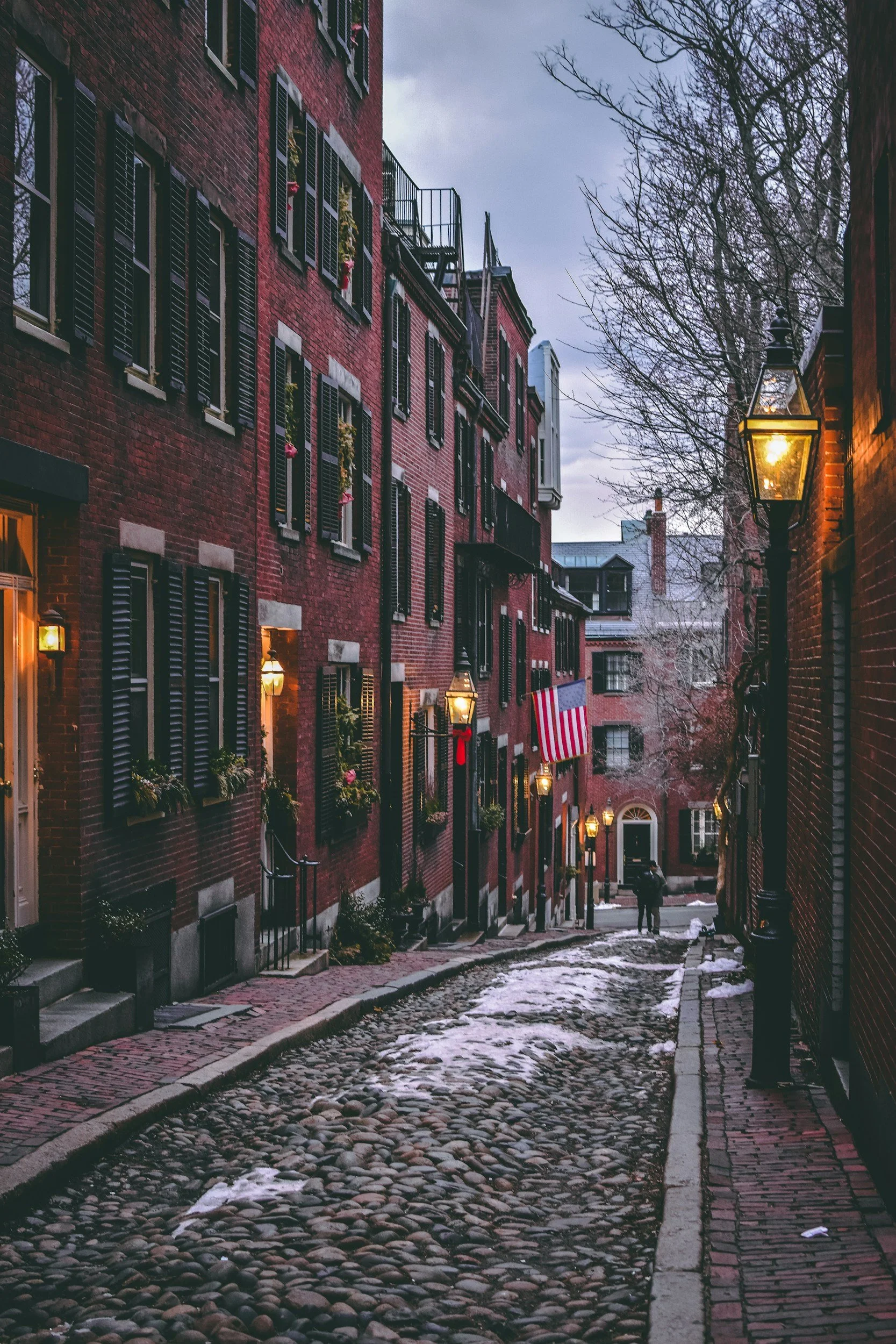 A cobblestone street lined with red brick buildings illuminated by wall-mounted lanterns and decorated with American flags and flower boxes, during dusk in a historic district.