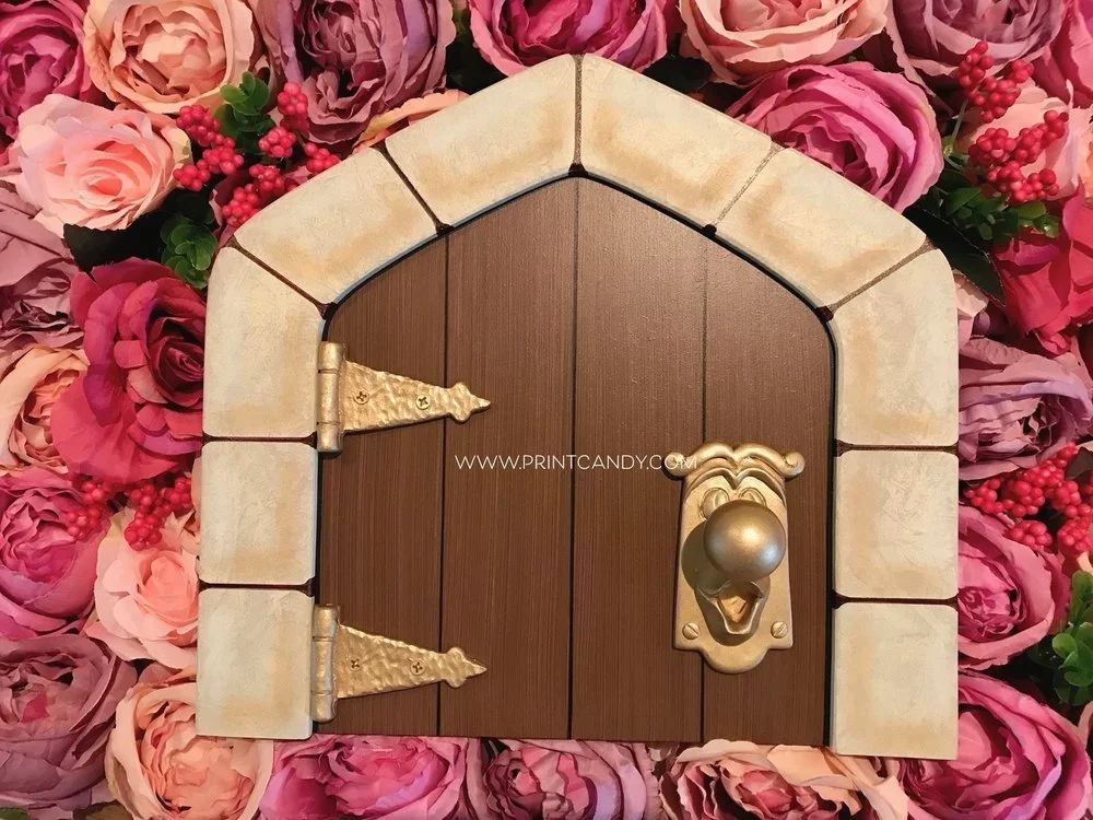 Miniature wooden door with brass hinges and a round brass door handle, surrounded by pink and purple roses with green leaves and red berries.