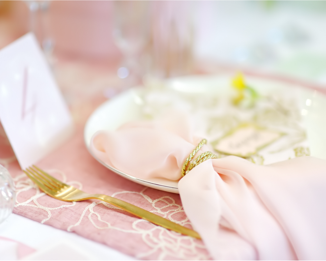 Elegant table setting with a gold fork, pink cloth napkin tied with a gold string on a white plate, and a blurred floral centerpiece, at a pastel-colored event or wedding.