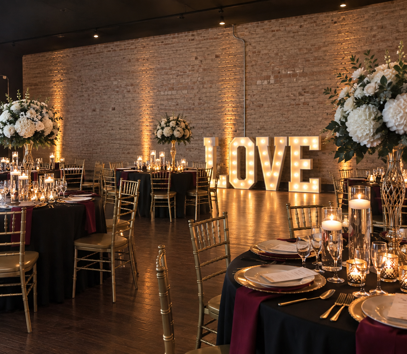 Wedding Venue At The Venue Taylor setup with decorated tables, tall floral centerpieces, candles, and large illuminated 'LOVE' sign against a brick wall.