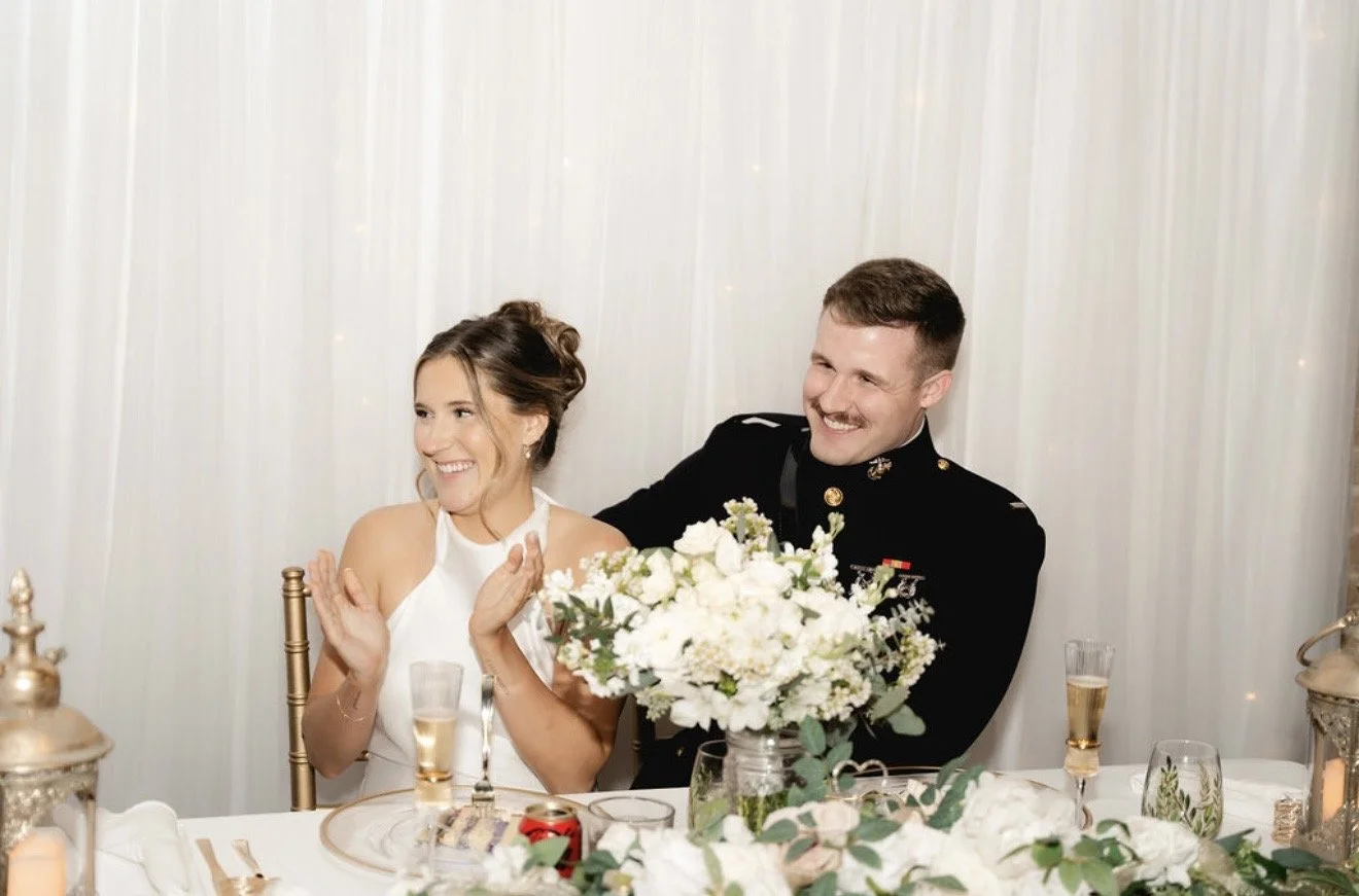 Happy bride and groom at wedding reception, laughing and clapping, seated at a decorated table with floral centerpiece, champagne glasses, and candles.