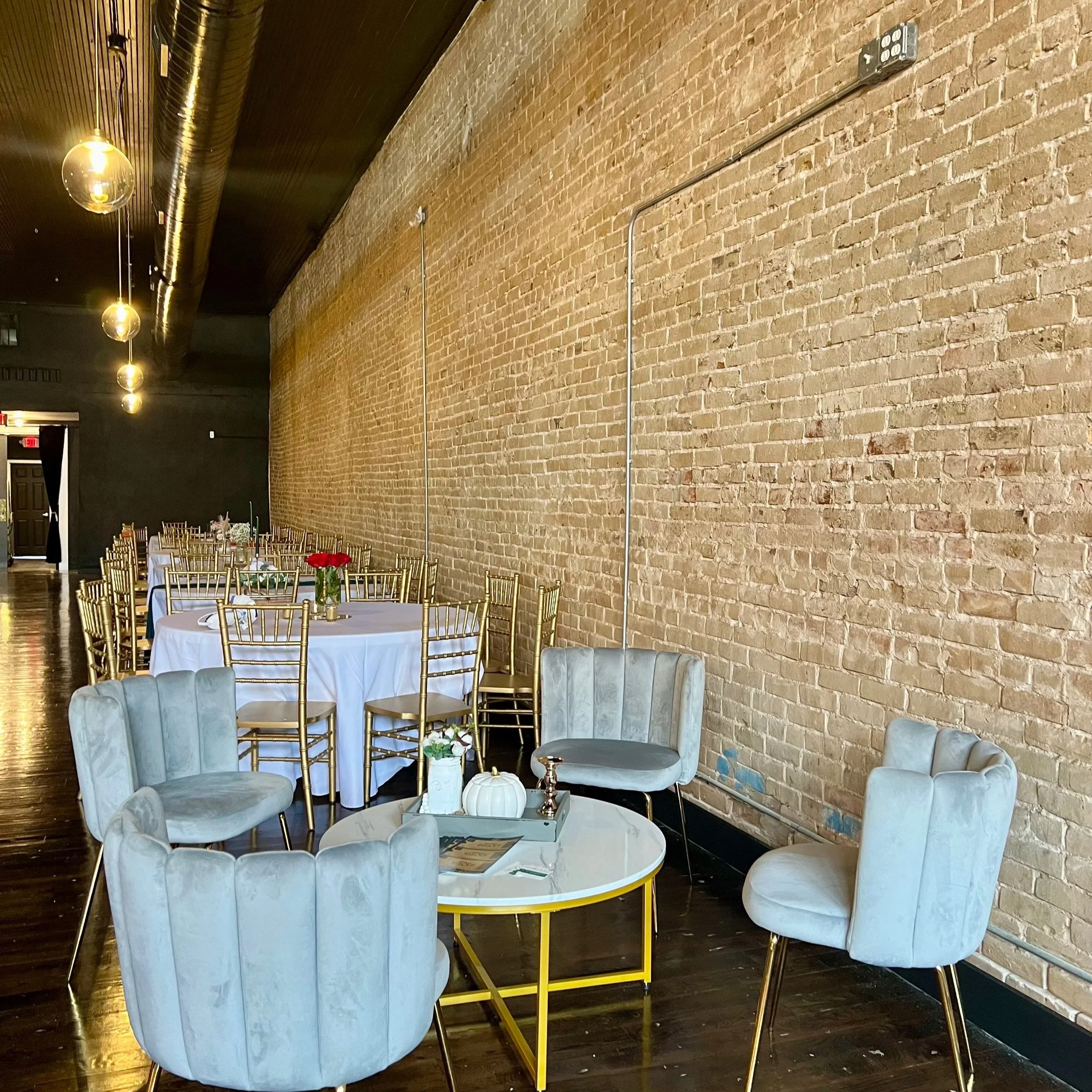 Indoor event space with brick wall, round tables with white tablecloths, gold chairs, and decorated centerpieces featuring flowers and pumpkins. Hanging lights and exposed ductwork are visible.