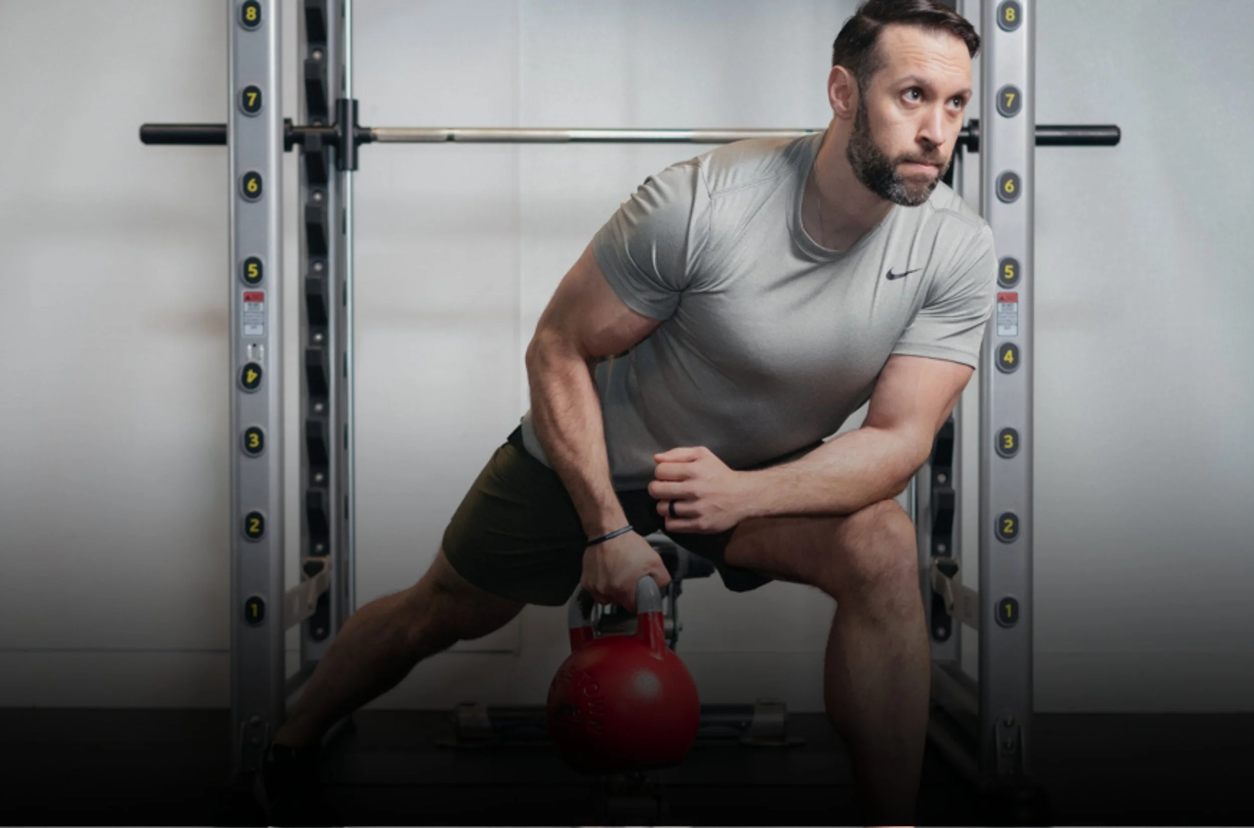 A man in workout clothes performing a side lunge with a kettlebell in his right hand inside a gym.