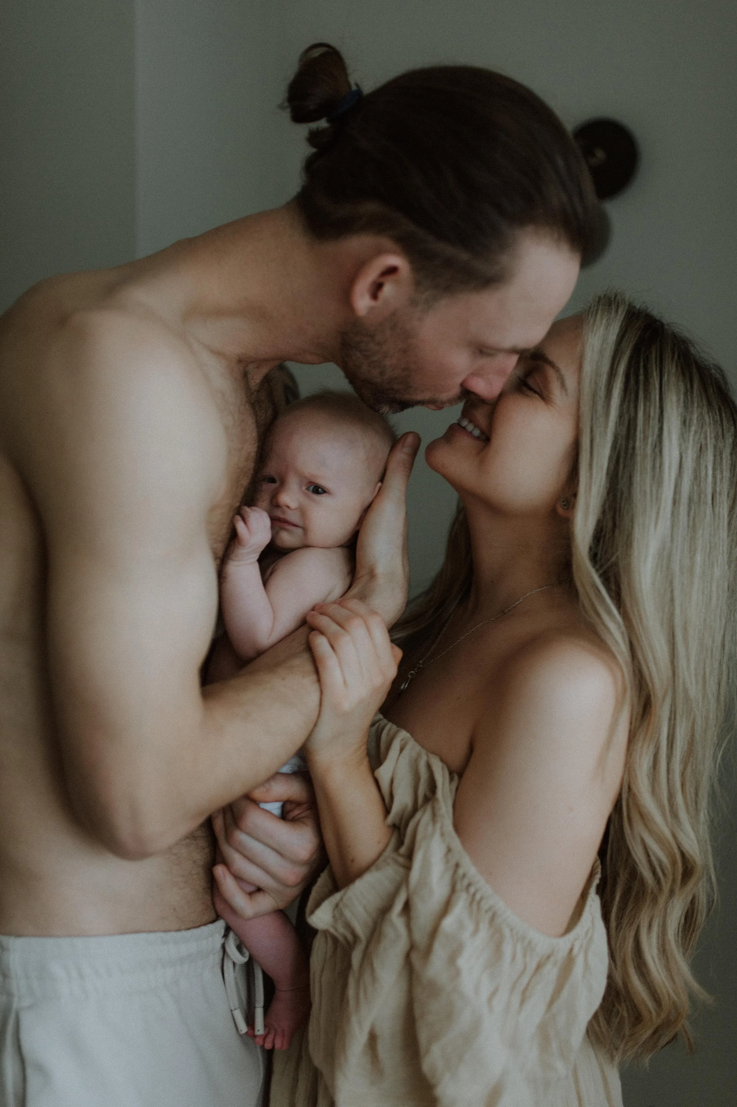 A shirtless man and a woman with long blonde hair hold a newborn baby close, touching foreheads and smiling, in a tender moment.