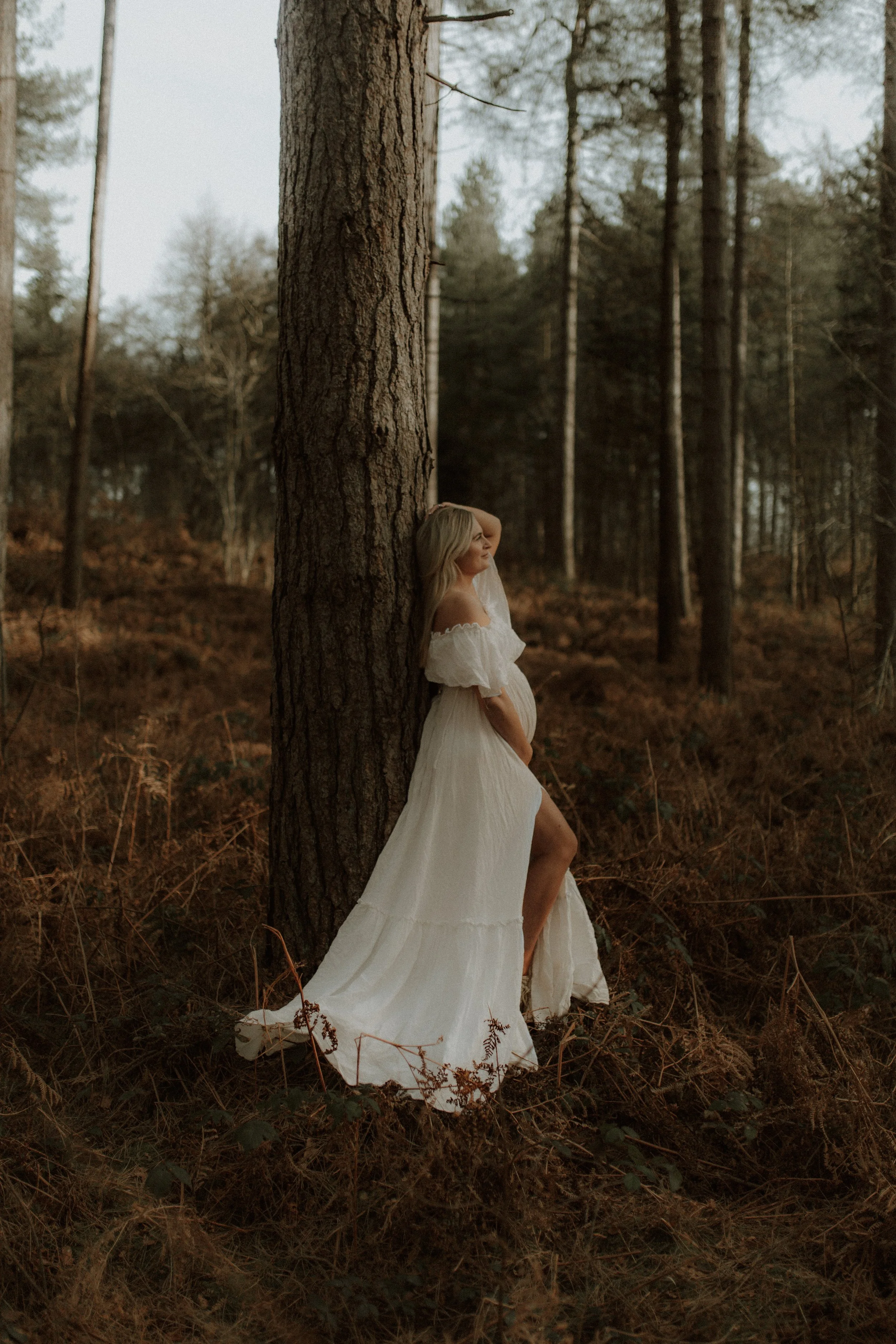 A pregnant woman in a white dress leaning against a large tree in a forest with tall trees and brown foliage.