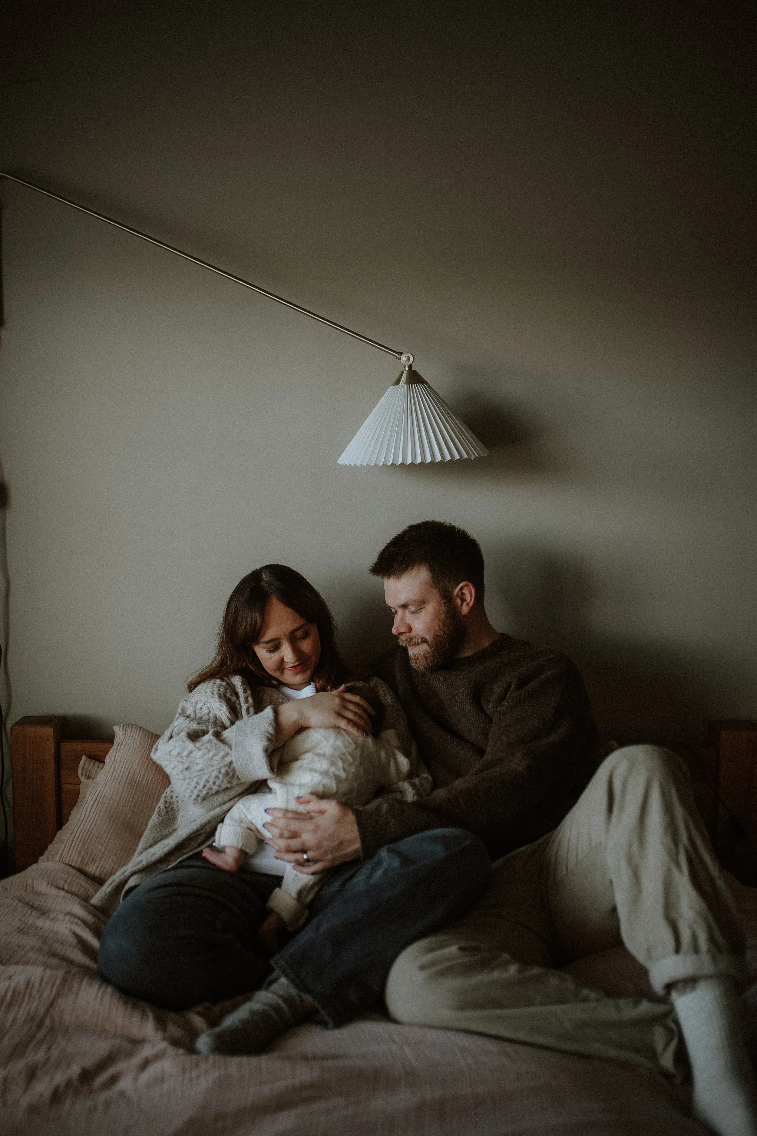 A family sitting on a bed; a woman and man holding a baby, with warm lighting and a hanging lamp above them.
