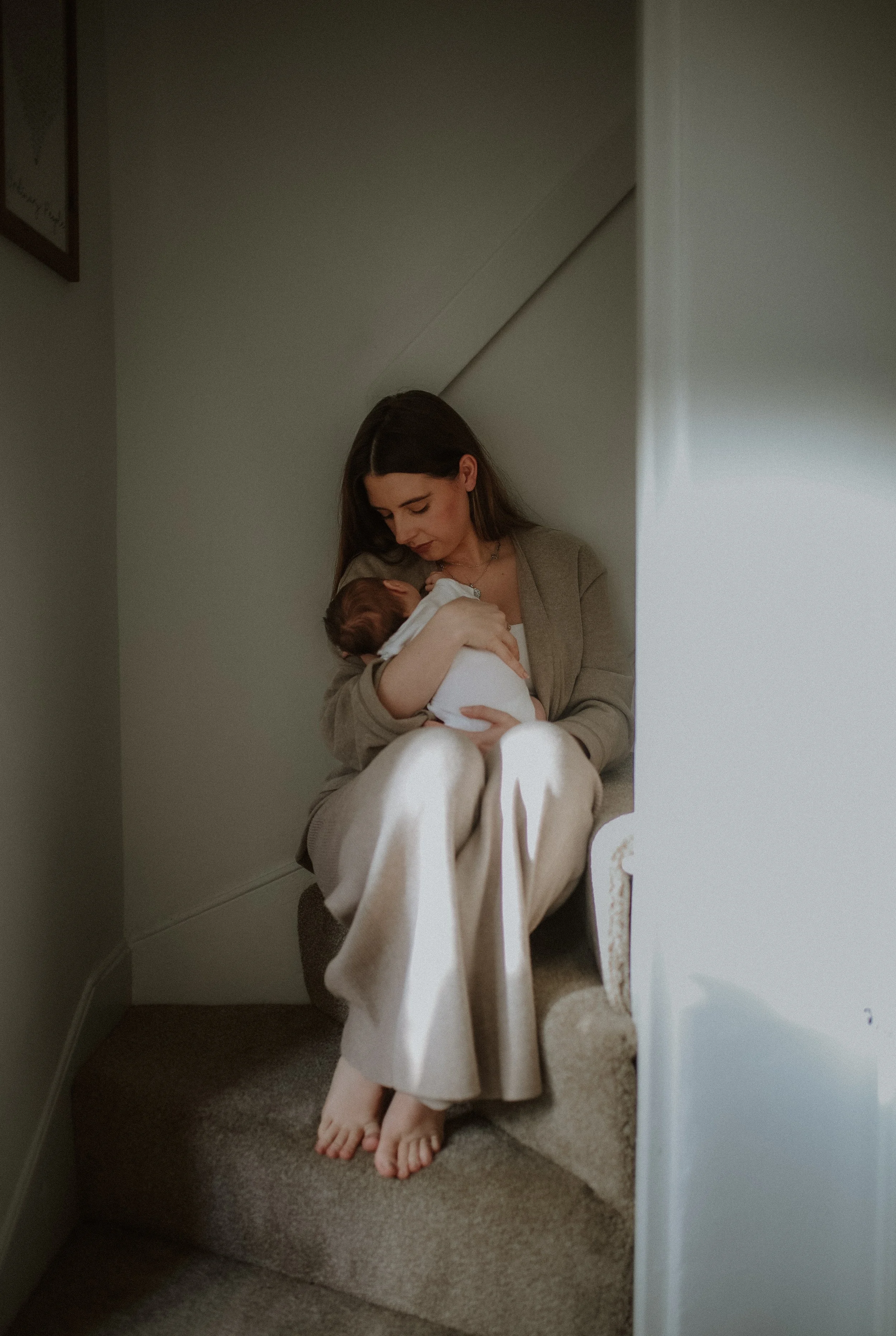 A woman sitting on stairs holding a newborn baby, looking down at the baby with affection in a softly lit indoor setting.