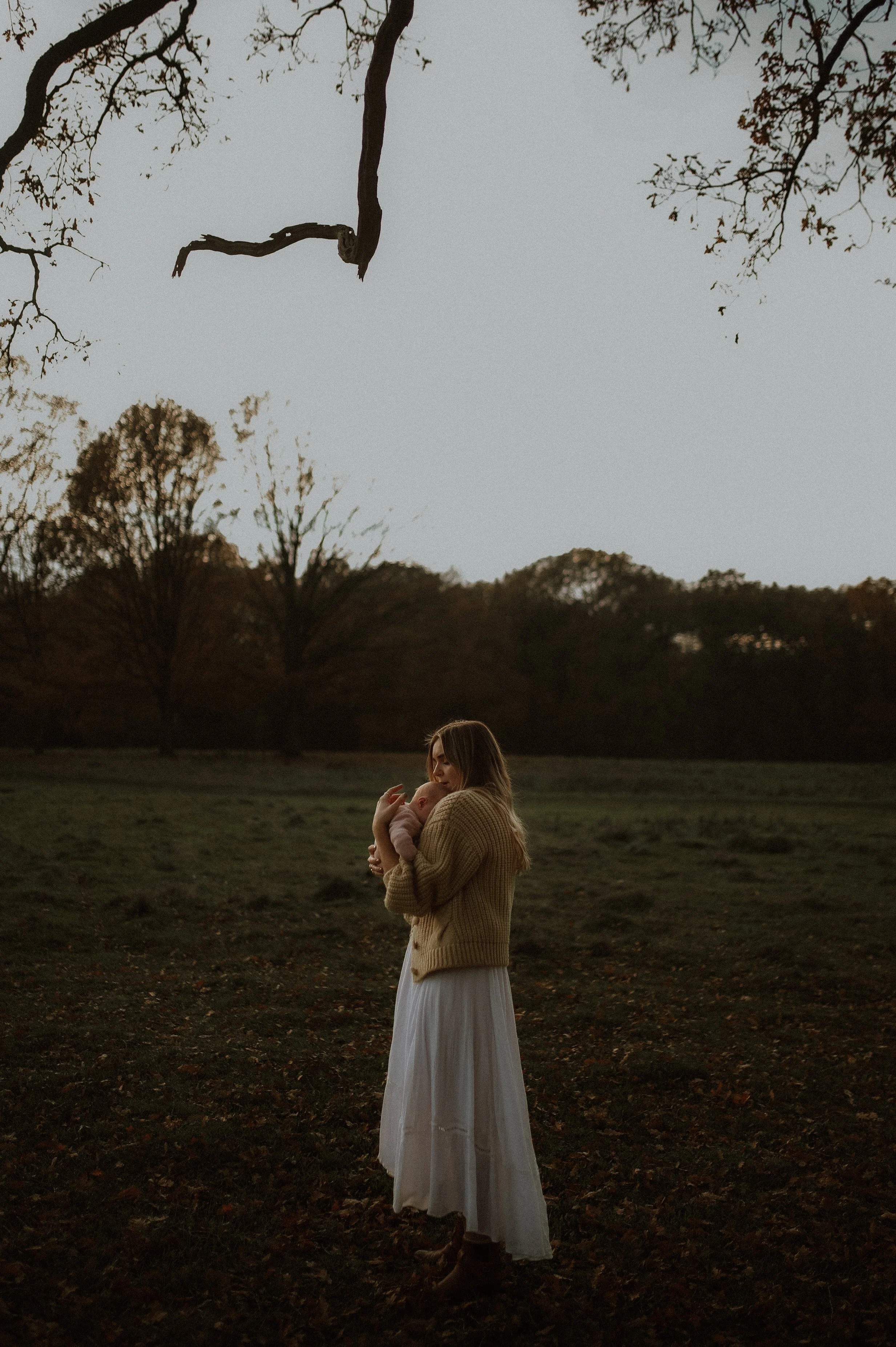 A woman holding a baby outdoors during sunset or dusk in a park with trees in the background.