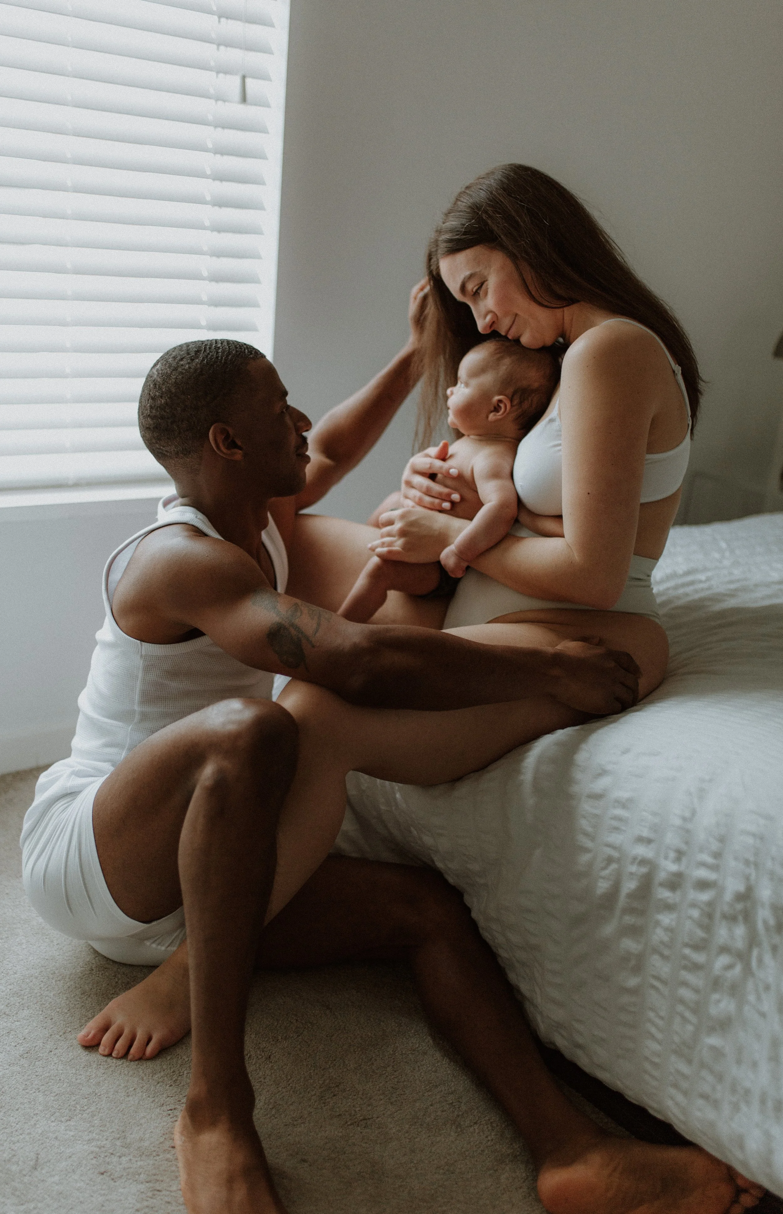 A diverse family of three on a bed, with the mother holding a baby, father kneeling, and all smiling at each other.