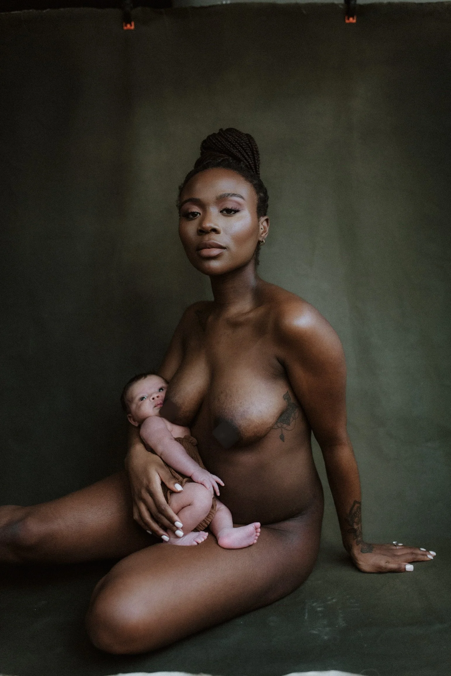 A topless woman with tattoos holding a newborn baby against a dark green background.