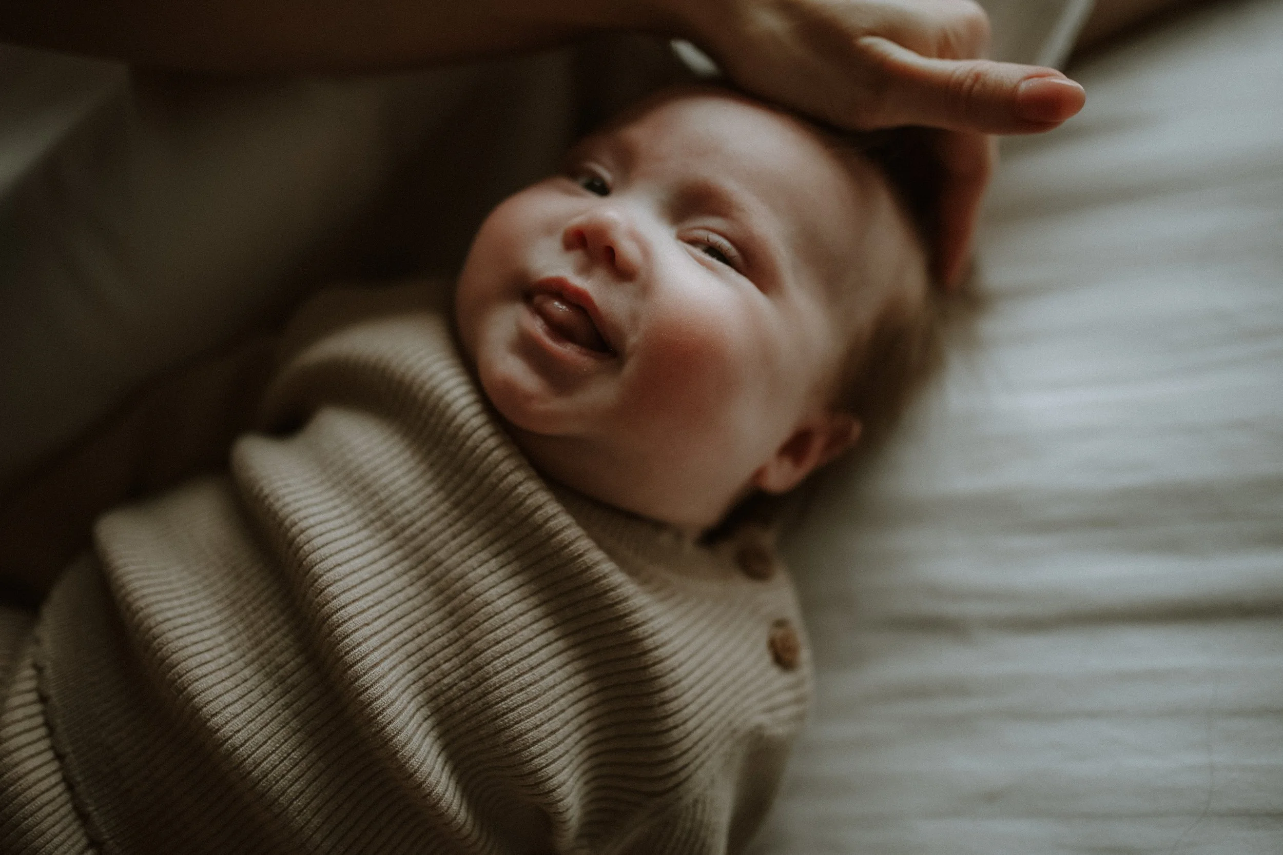 A baby with a happy expression lying on a bed with a neutral pillow, looking at the camera, and being gently touched on the head by an adult.