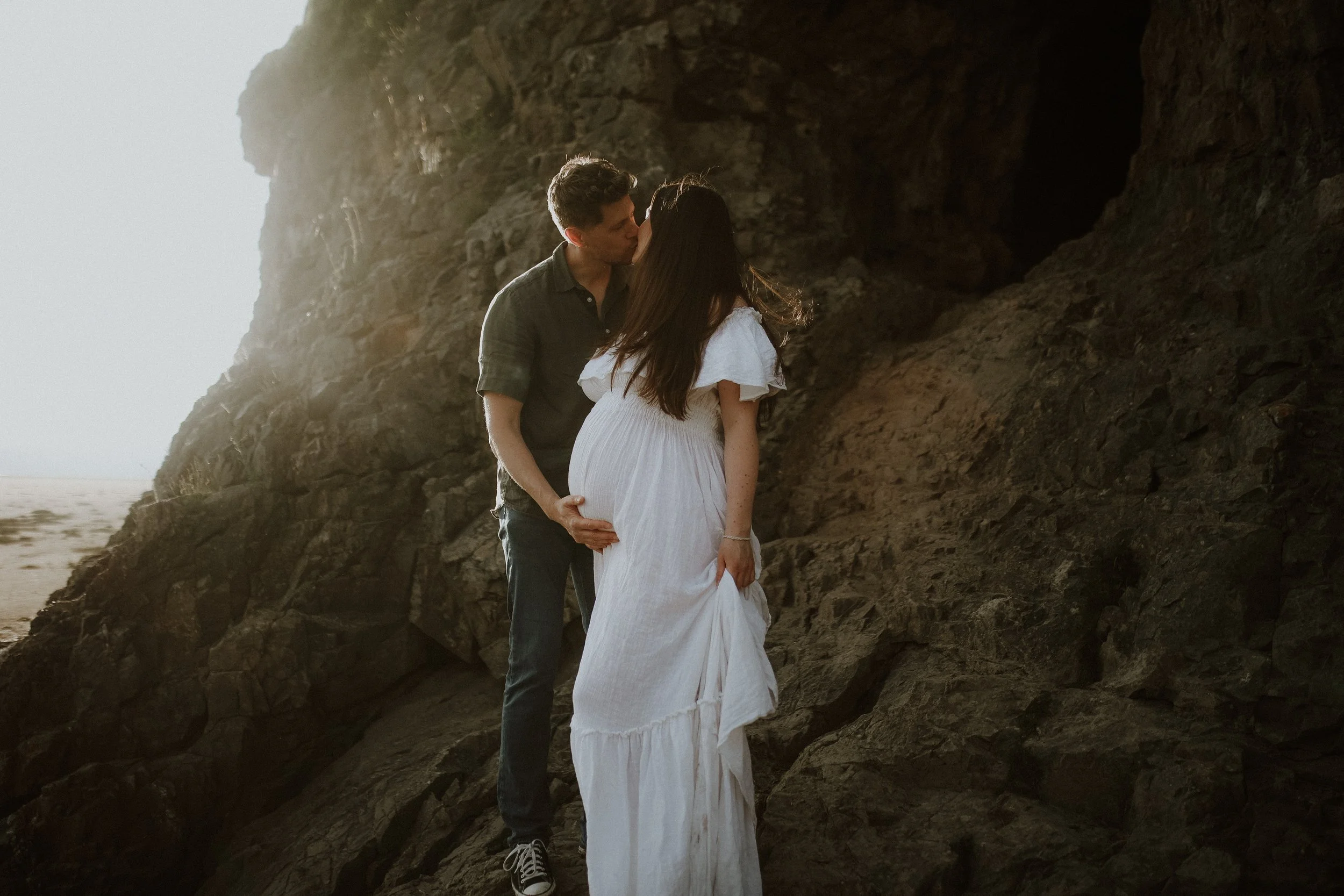 A couple kissing on a rocky beach, the pregnant woman is wearing a white dress, and the man is in a green shirt and jeans.