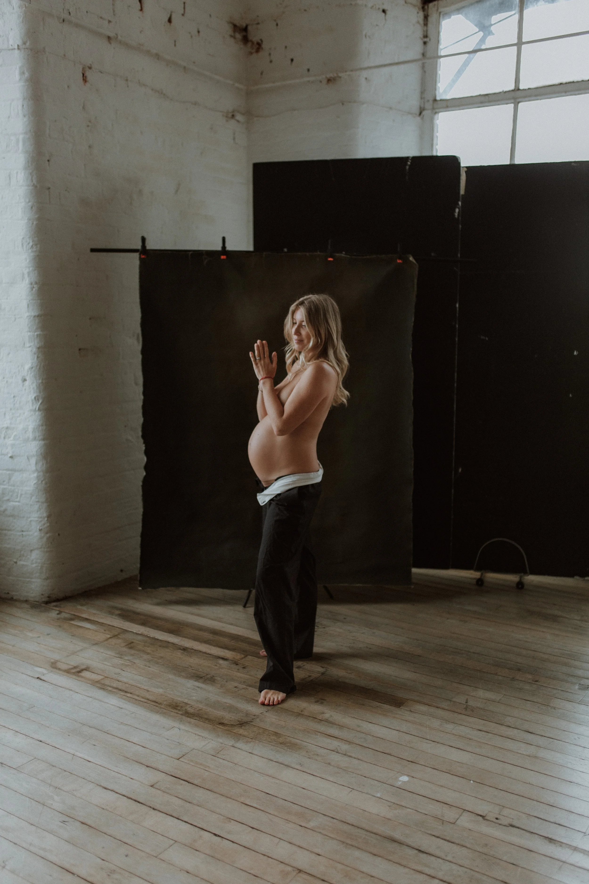 A pregnant woman standing and posing indoors in front of a black backdrop, with her hands together, wearing black pants and a white shirt pulled down to show her belly, in natural light from a large window.