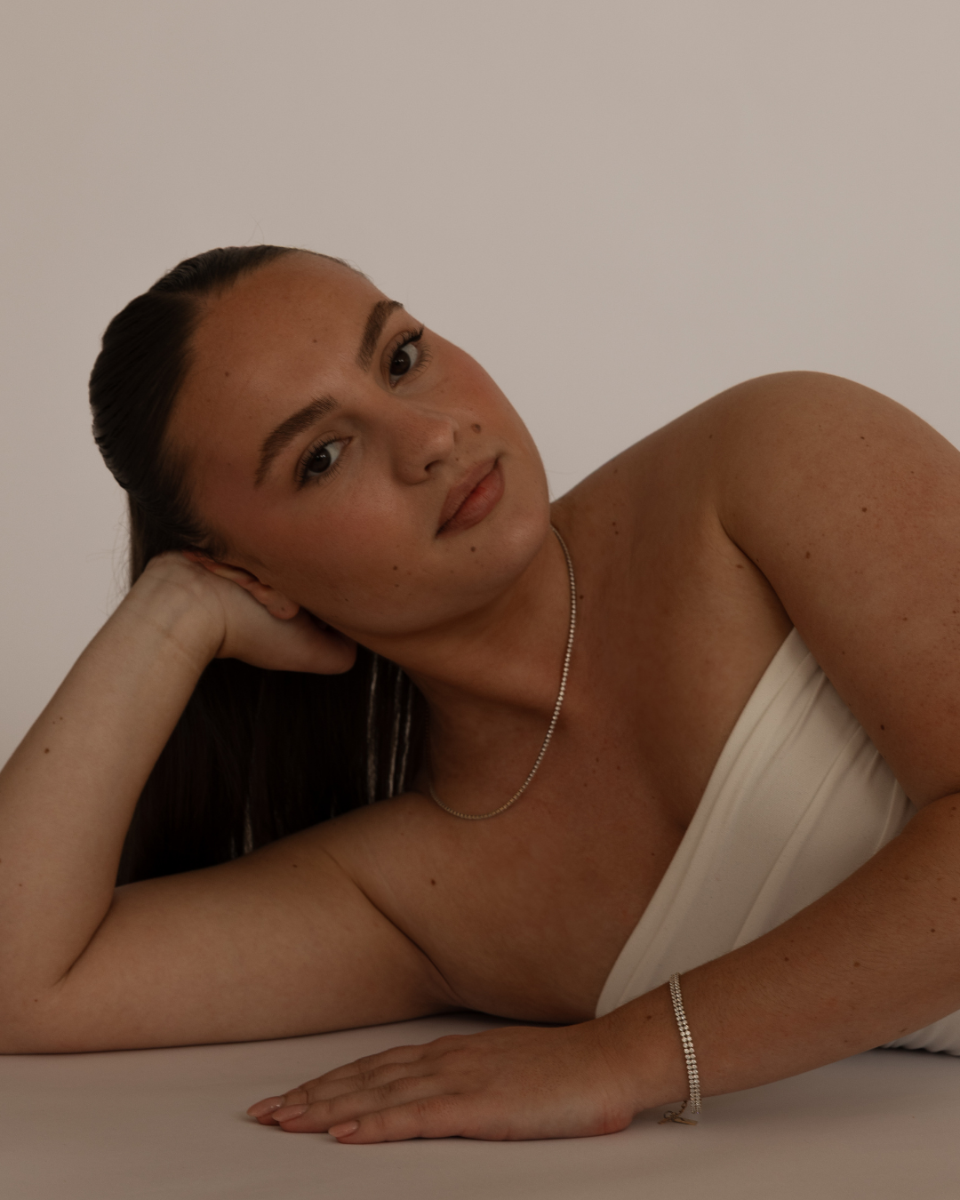 A woman with dark hair styled back, lying on her side with her head resting on her hand, wearing a strapless top and jewelry, against a plain background.