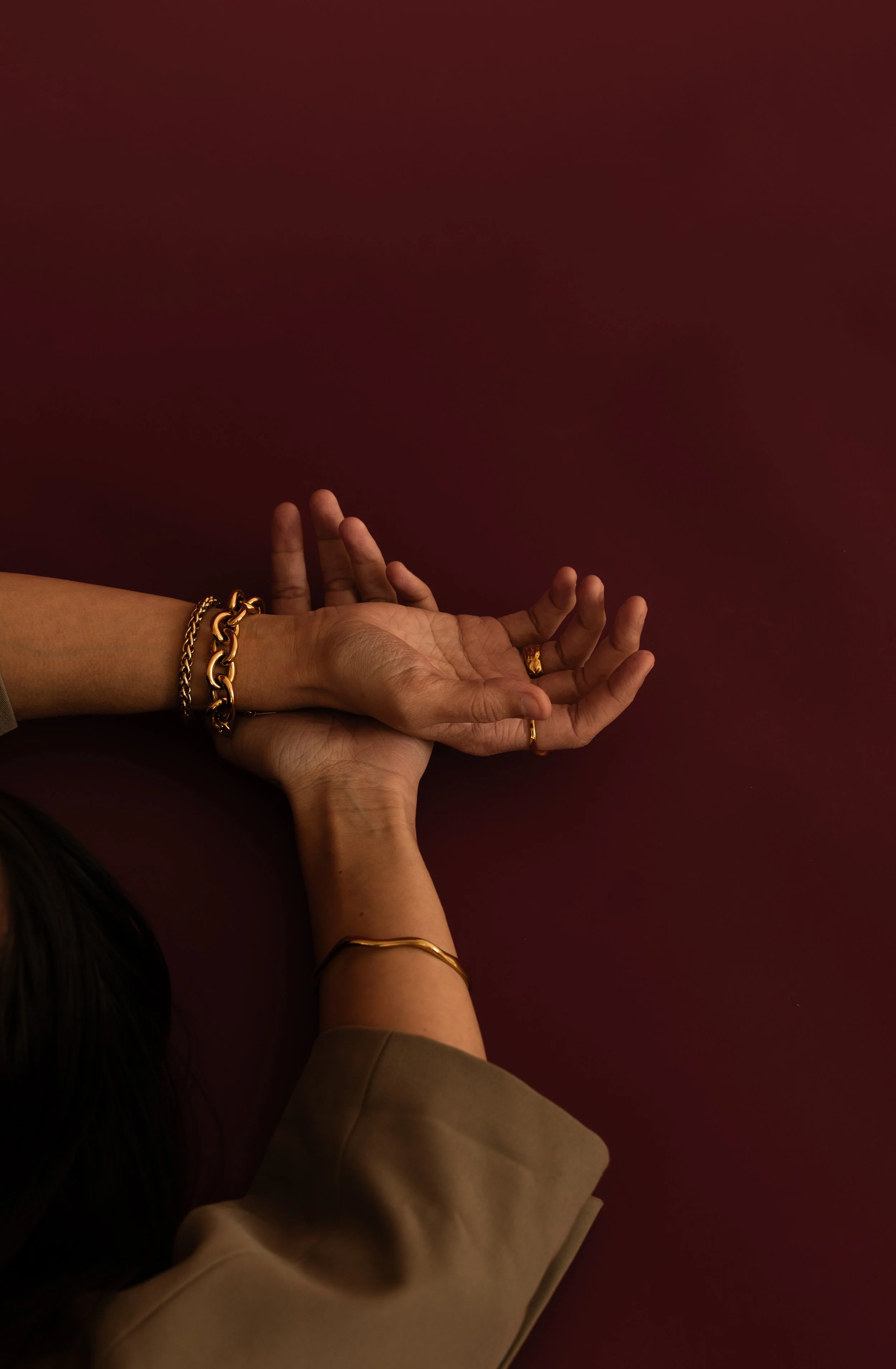 Two hands, one with gold jewelry, resting on a maroon surface, partially covered by a beige sleeve.