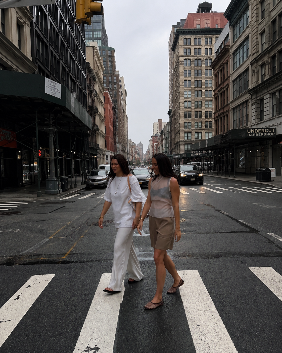 Two women crossing a city street on a crosswalk, with tall buildings and cars in the background on an overcast day.