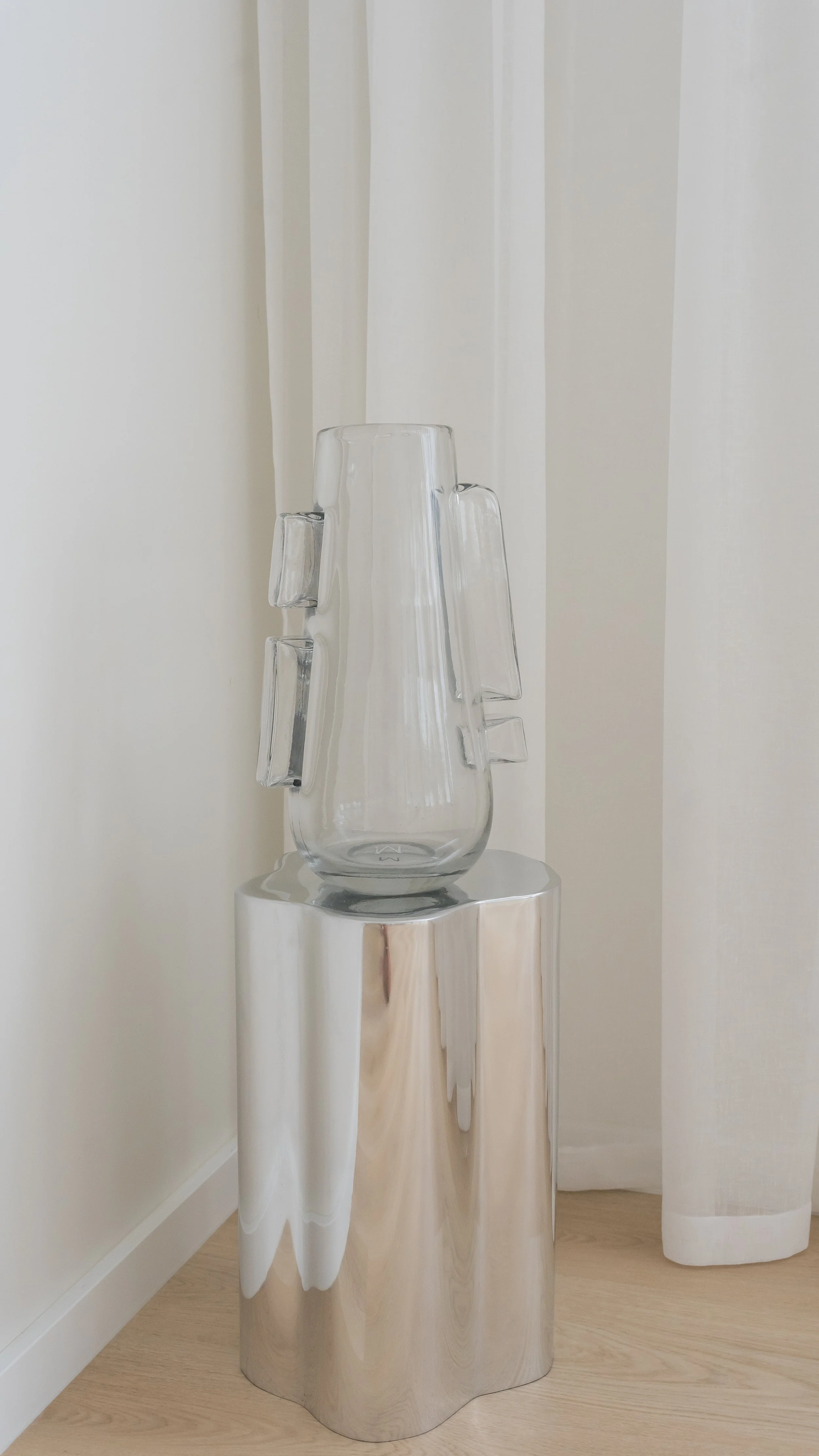 A clear glass vase with four extra glass handles attached on the side, placed on a reflective silver pedestal.