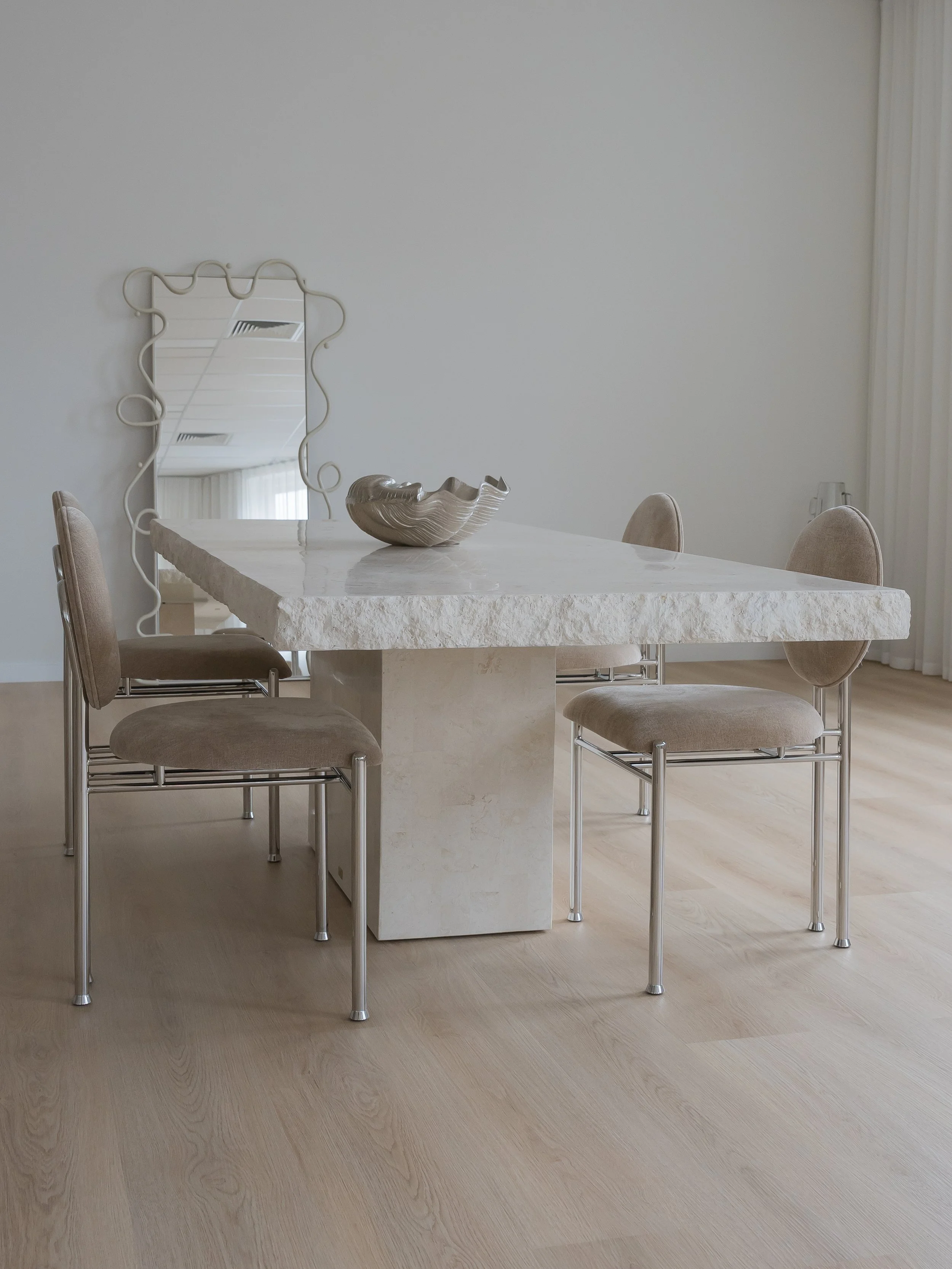Minimalist dining room with a rectangular stone table and six beige upholstered chairs with metal legs. A decorative ceramic bowl is on the table, and a uniquely shaped mirror leans against the wall in the background.