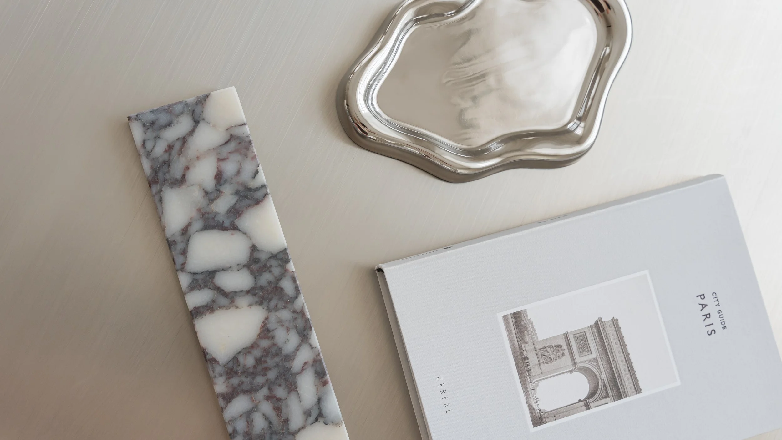 A marble tissue box cover, a silver decorative tray, and a Paris city guidebook on a light-colored wooden surface.