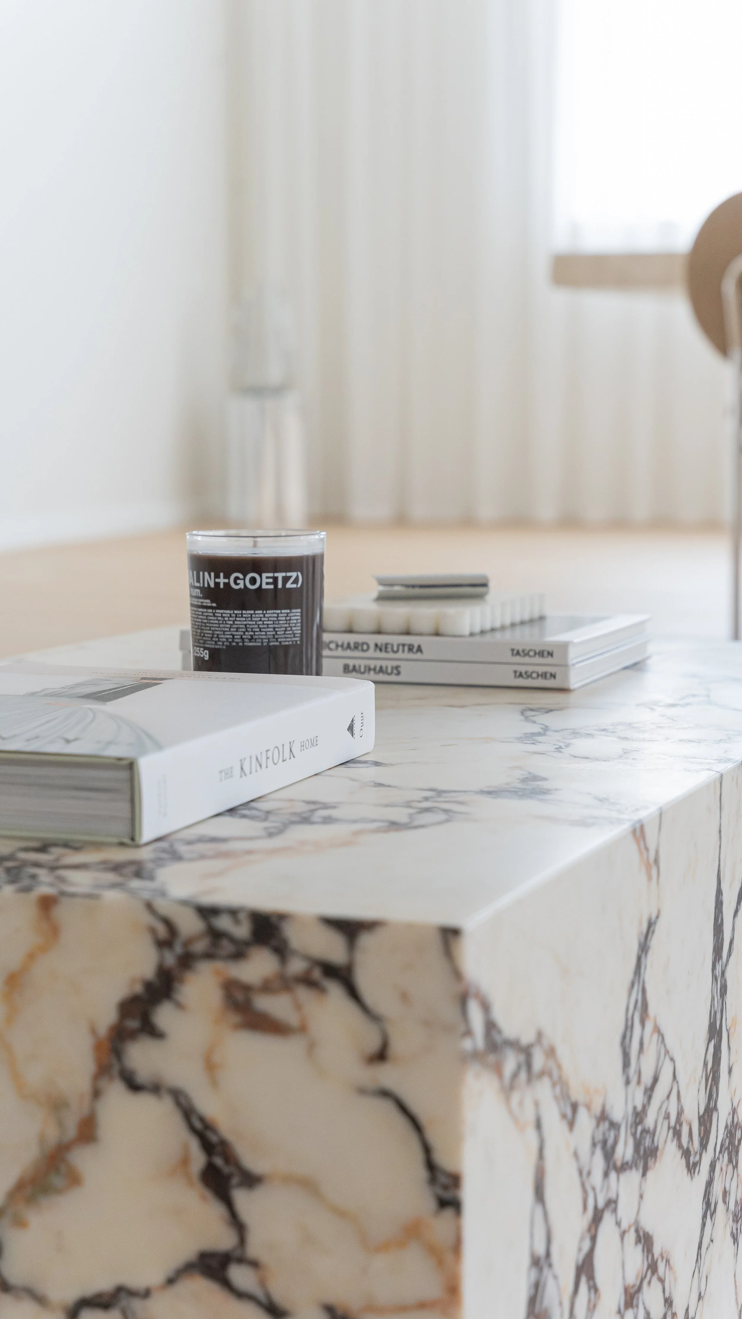 A marble table with a candle, a book titled "The Kinfolk Home," and a stack of art books with titles "Richard Neutra" and "Bauhaus" in a bright, minimalist room.