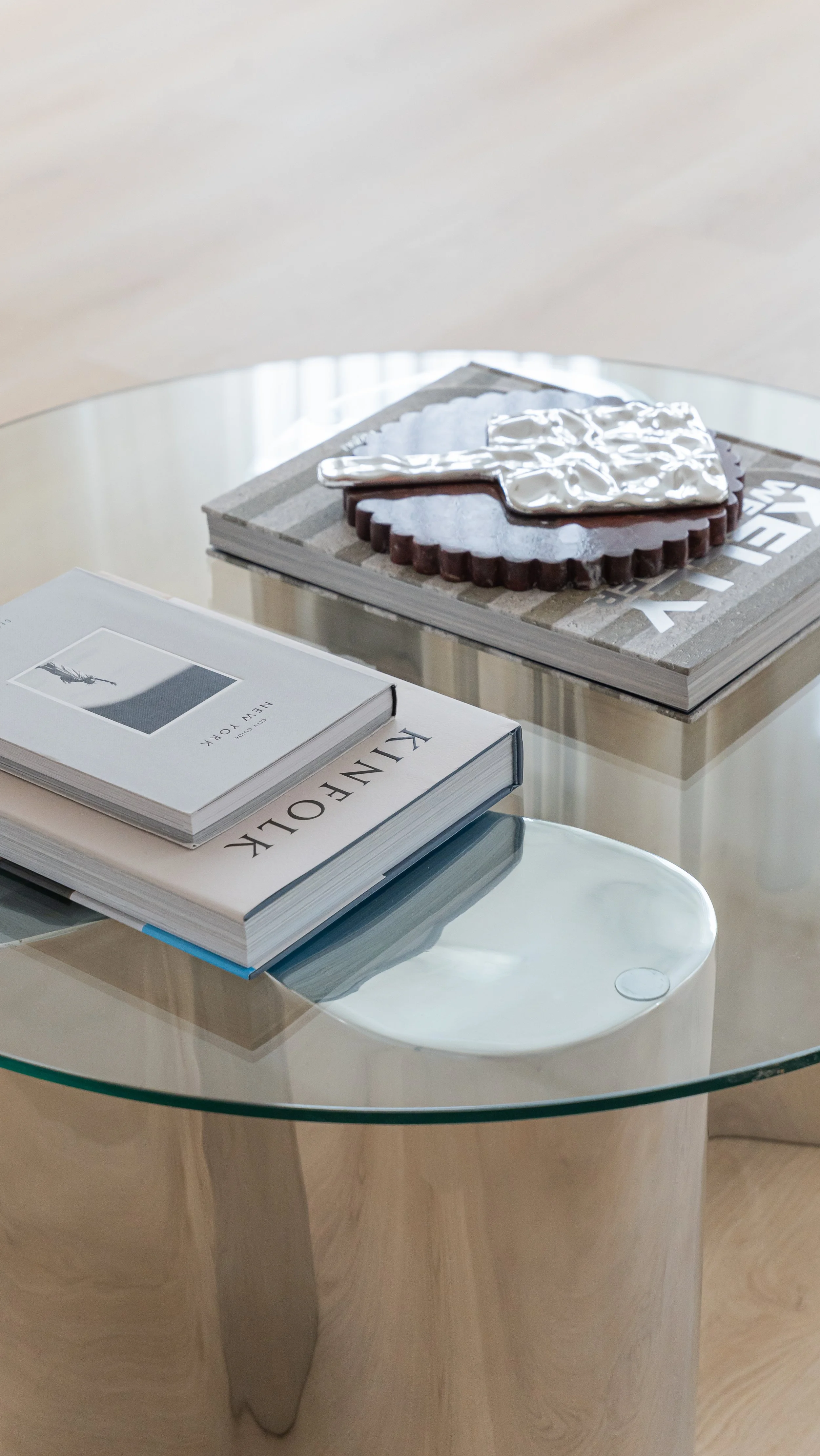 A glass coffee table with magazines, one titled 'KINFOLK' and the other with a black and white photo, and a decorative object on top of a book, next to a piece of artwork with shaped metallic and textured elements.