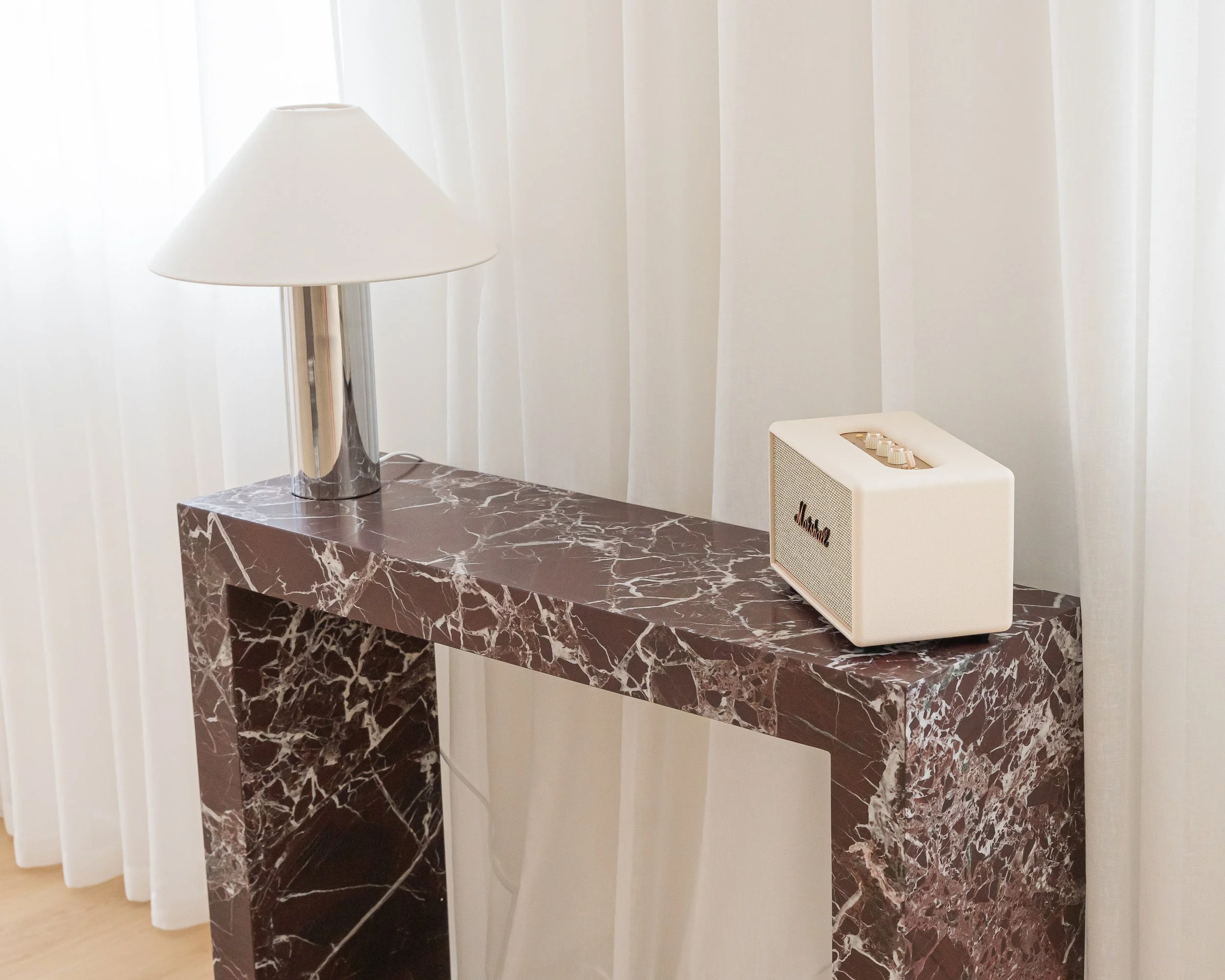 A marble console table with a white lamp and a small white speaker on top, positioned against white curtains.