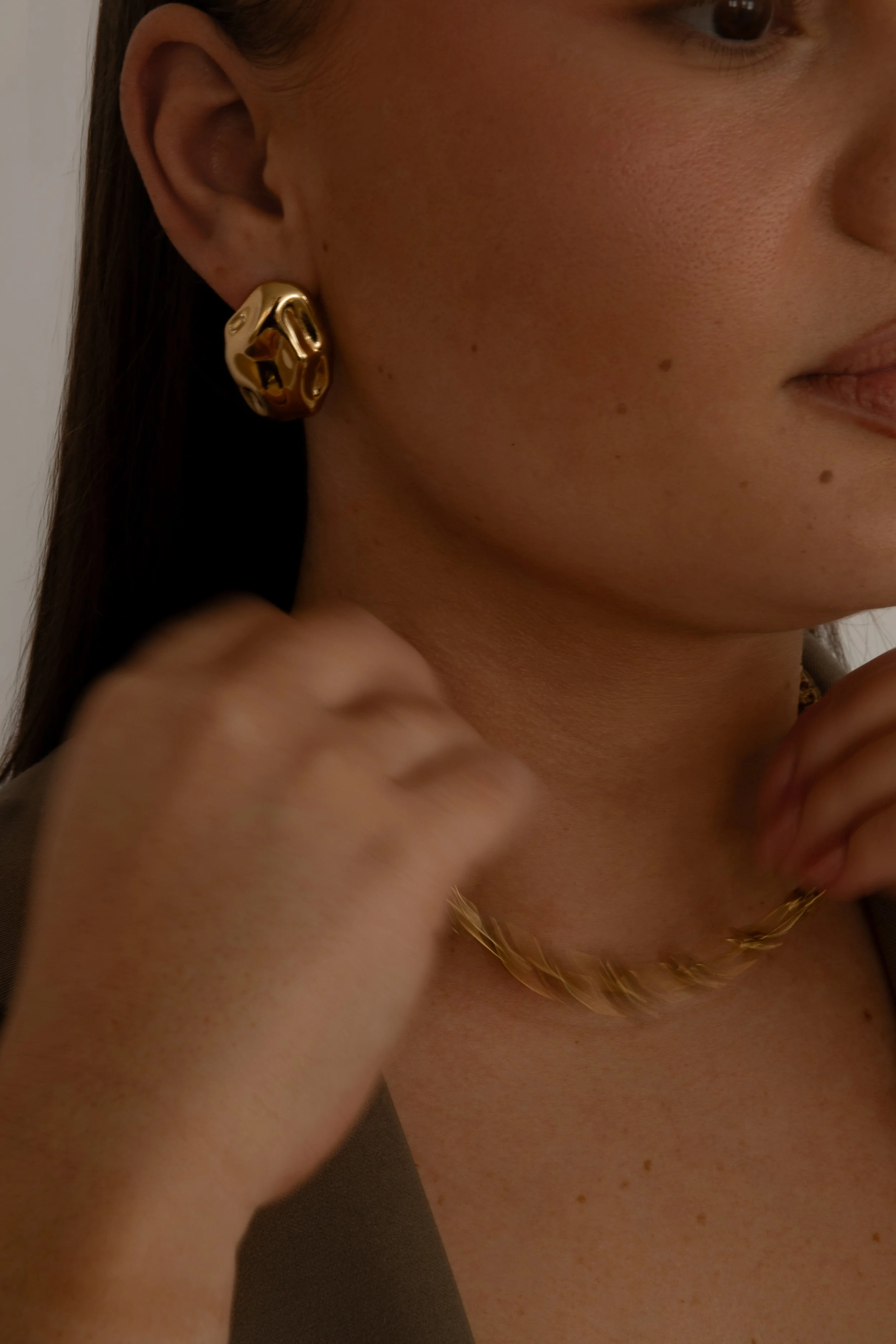 Close-up of a woman's face focusing on her ear with a gold statement earring, part of her lips, and a gold necklace visible.