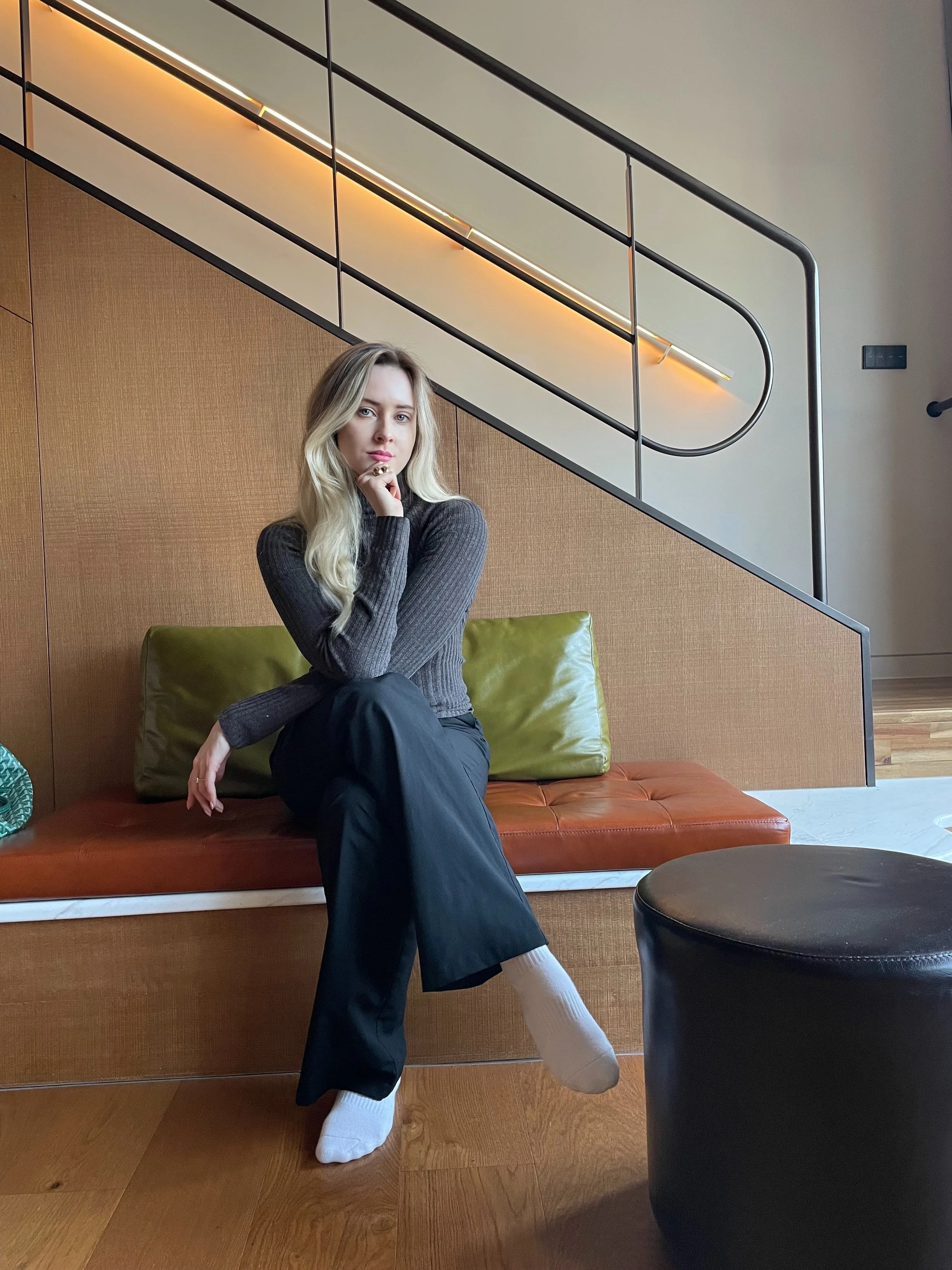 Nuori nainen istuu erilaisella penkillä sisätiloissa, taustalla on puupanelointi ja rappuset, joilla on metallikaiteet ja lämmin valaistus.