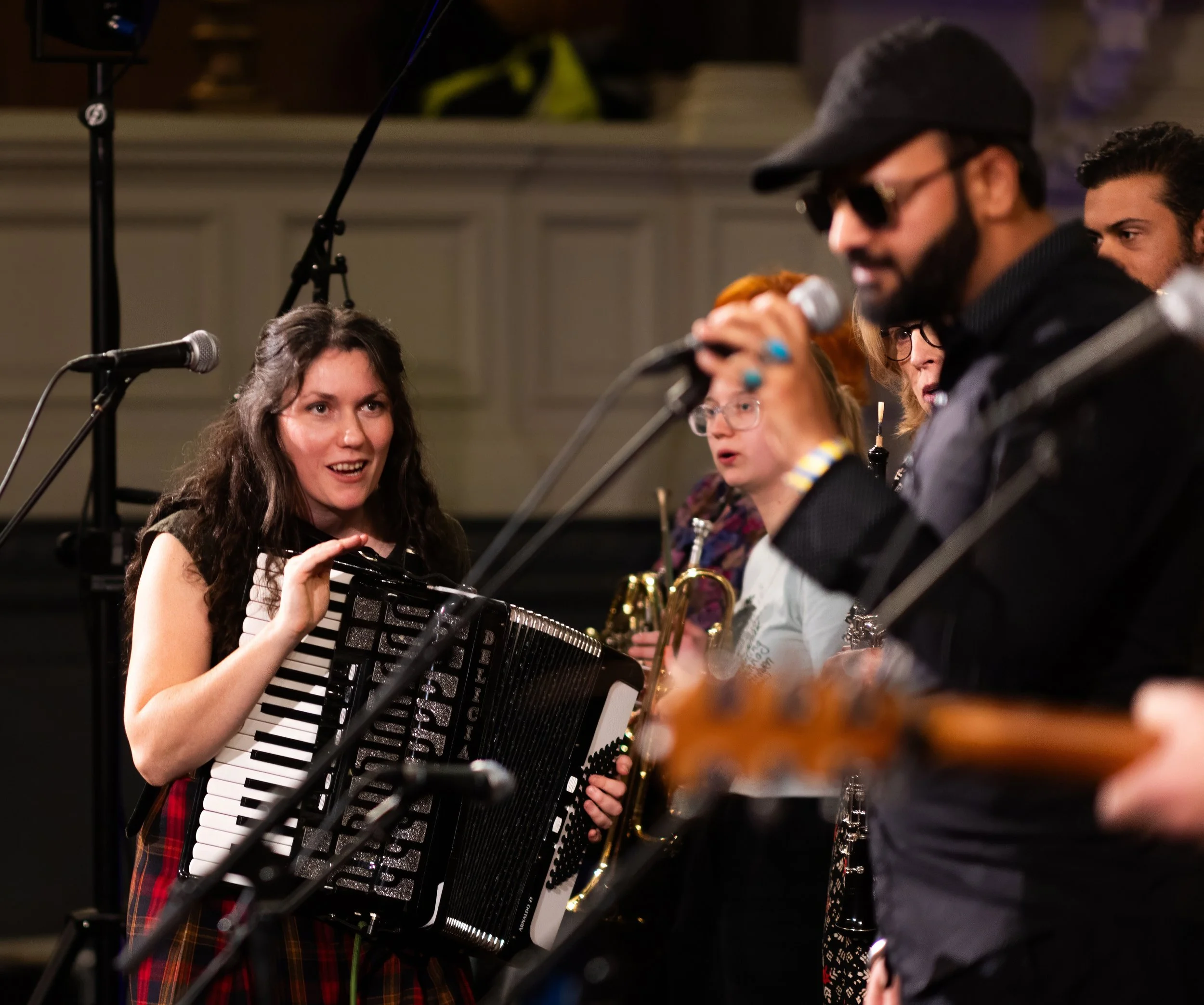 A group of people playing music on stage. There is a woman with an accordion, a man singing into a microphone and various other instruments including guitars and brass