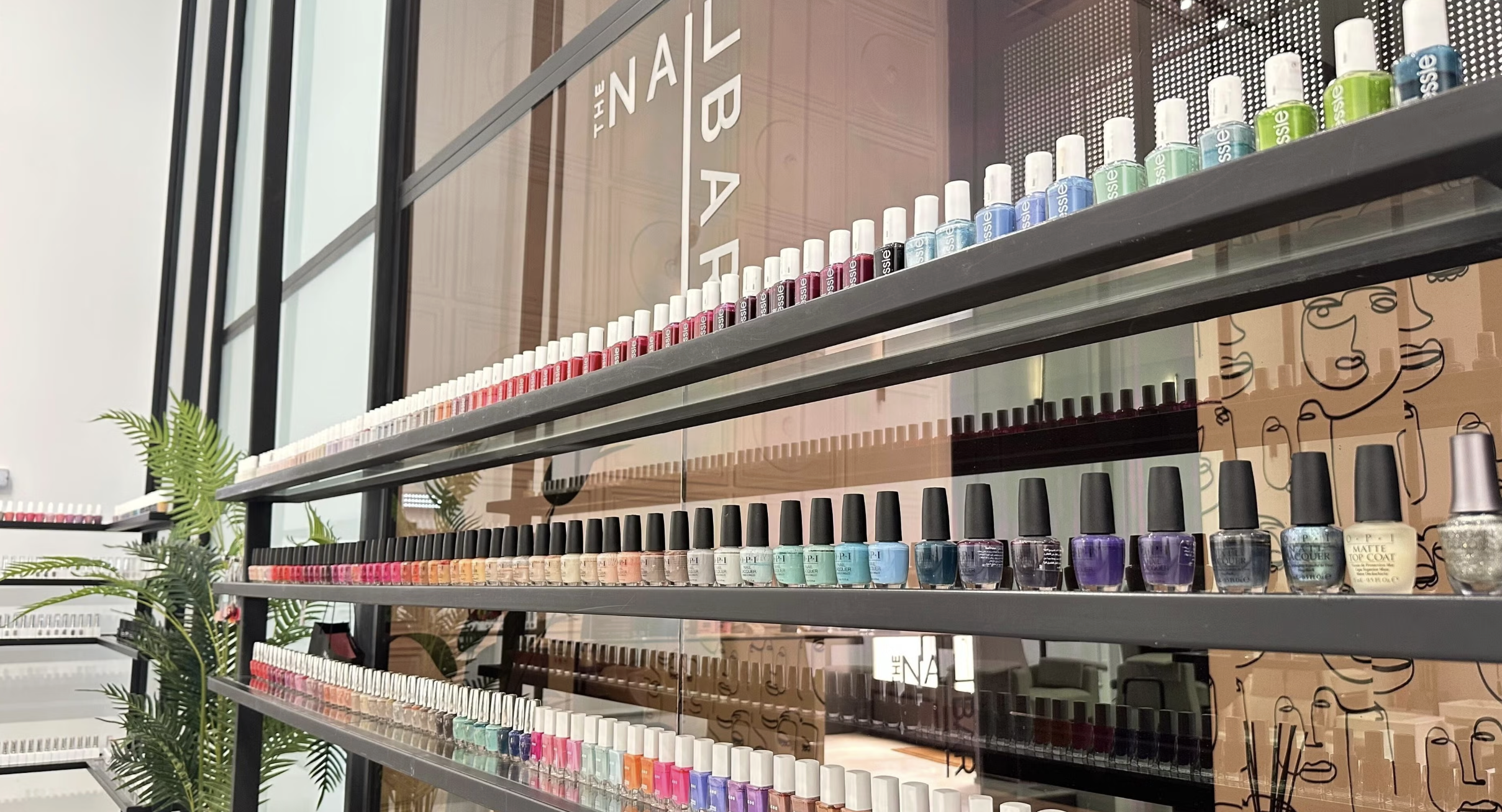 A display of assorted nail polish bottles in various colors on metal shelves in a nail salon, with decorative plants and a mirror reflecting part of the interior.