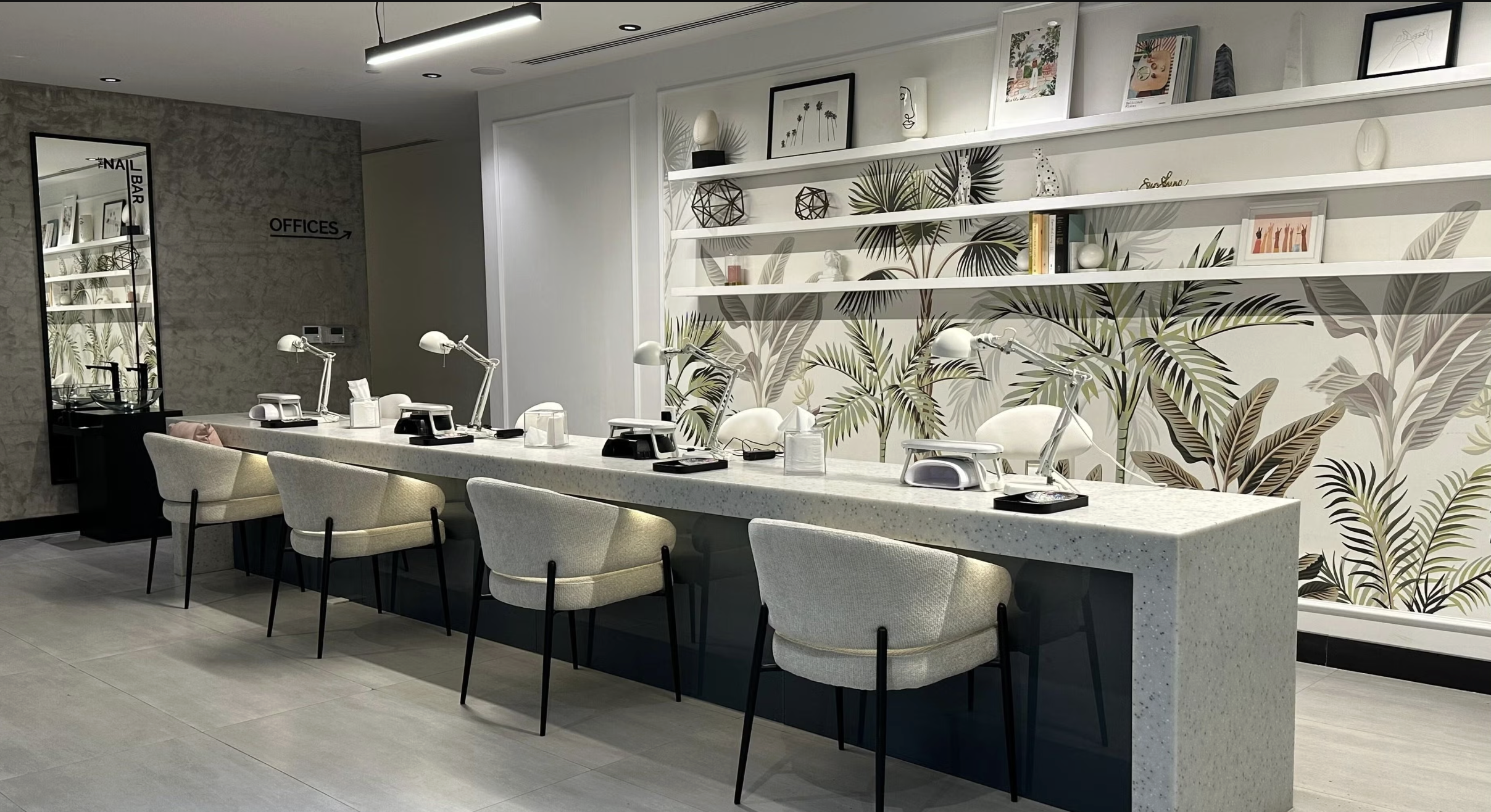 Modern nail salon with a long, speckled white manicure station and six cream-colored chairs. On the station, there are white desk lamps, manicure tools, and tissues. Behind the station, a decorative wall with tropical leaf wallpaper and white shelves displaying artwork, books, and decorative items. To the left, a mirror and a sign indicating the direction to the offices.
