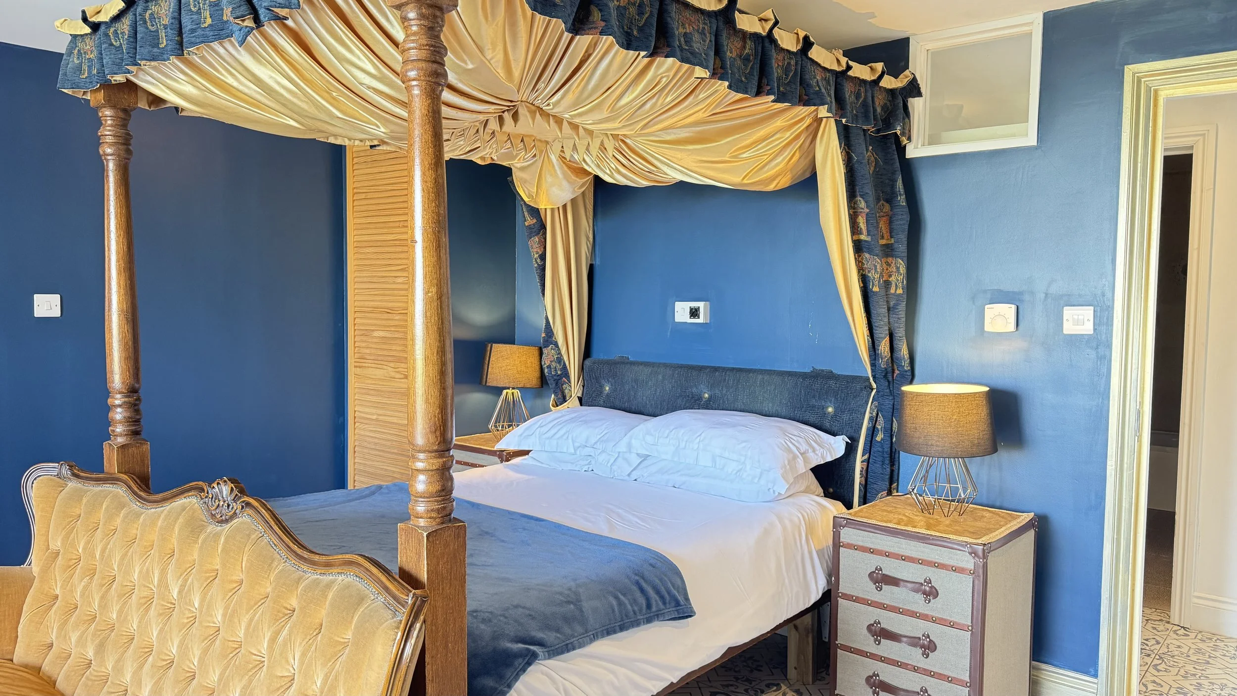 Bedroom with blue walls, a four-poster bed with golden drapes, a nightstand with a lamp, and a brown upholstered chair. Ocean view