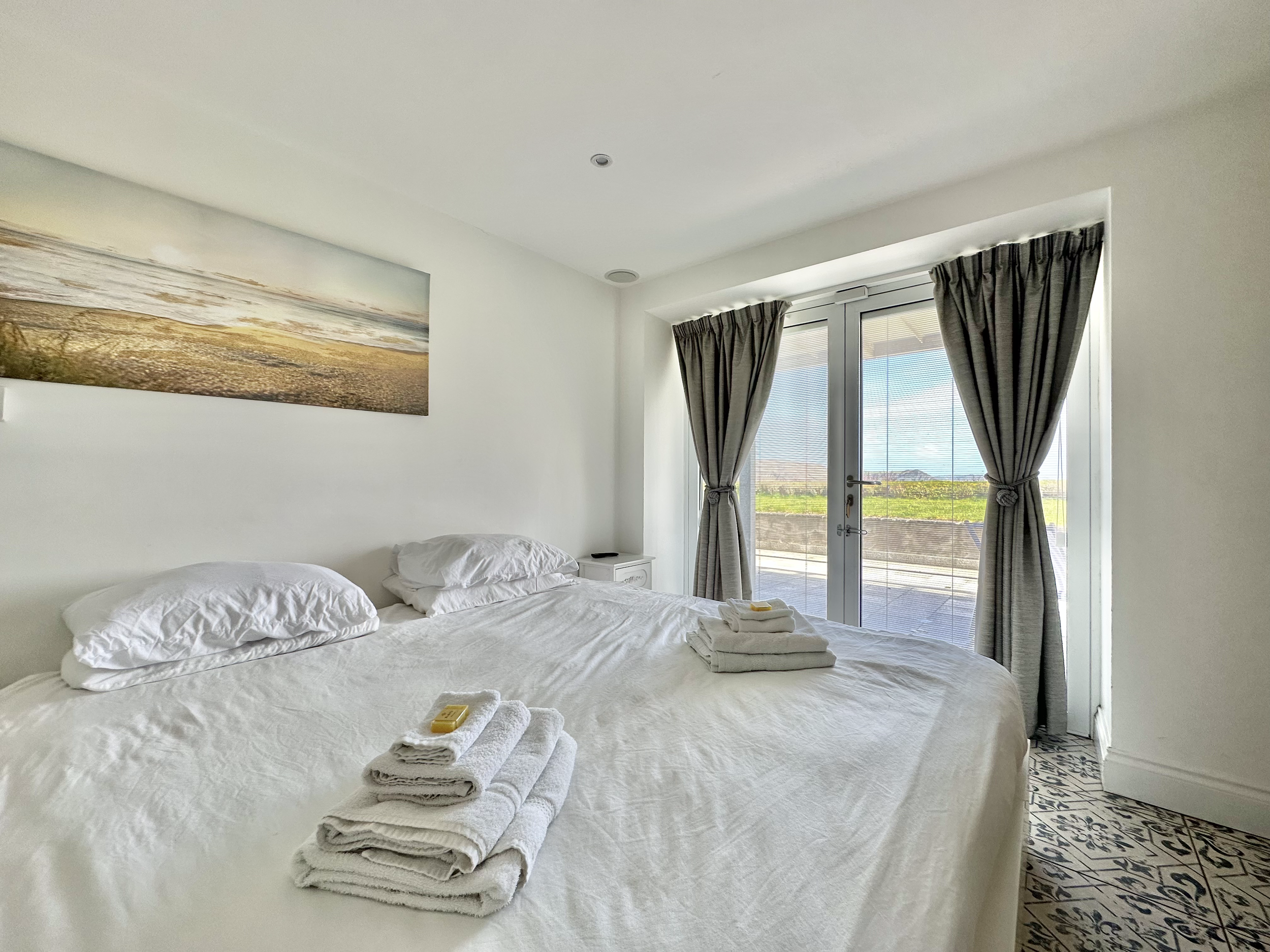 A bright and minimalistic bedroom with a large bed covered in white linens and neatly folded towels on top. The room features a landscape painting on the wall and a set of glass doors with dark curtains, offering a view of the ocean right outside.