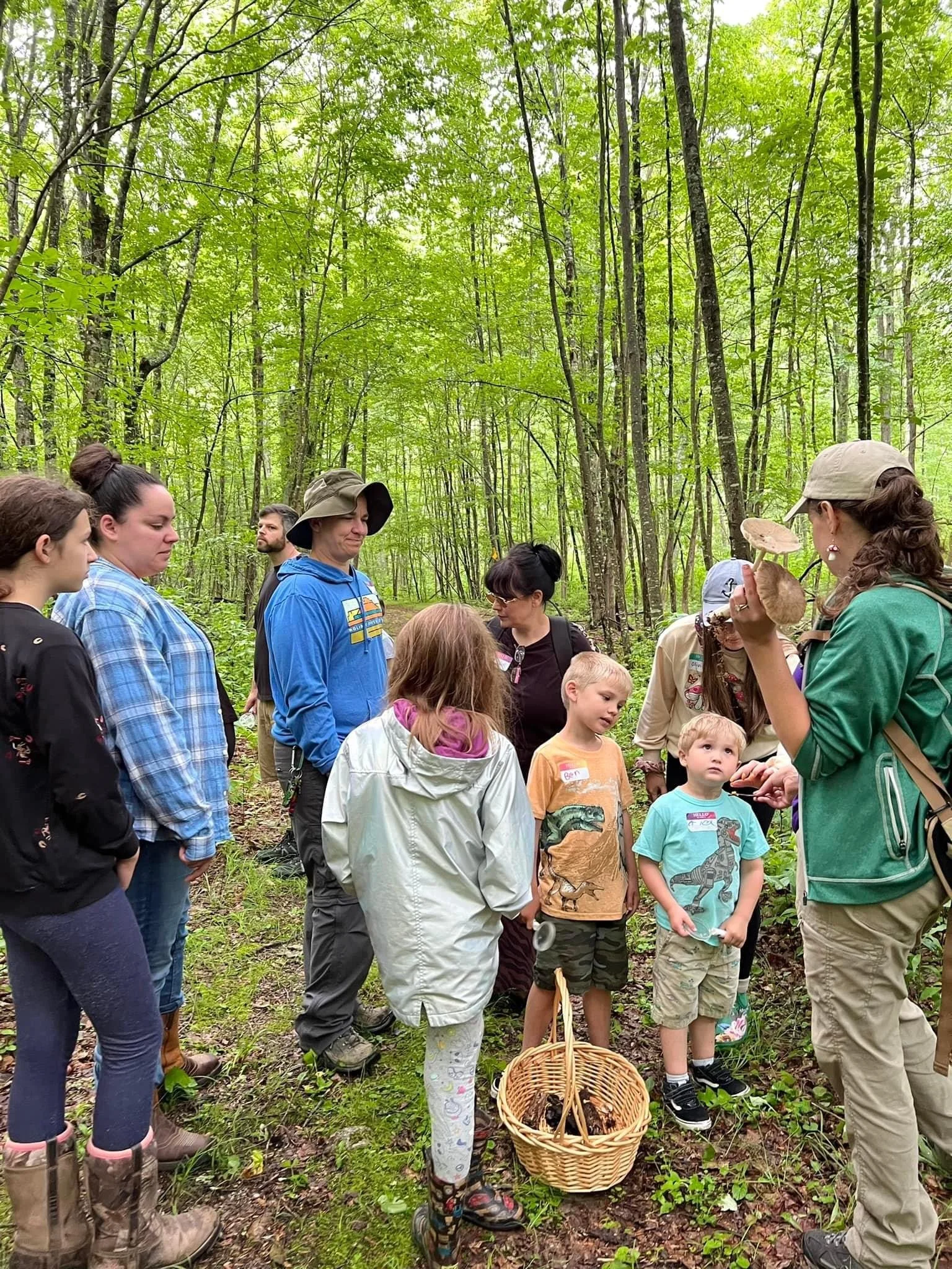 A group of children and adults gathered in a forest, listening to a woman hold up mushrooms for discussion.