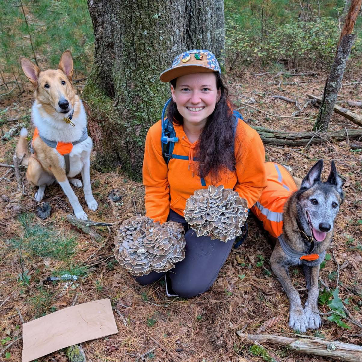 A woman smiling in an orange jacket with two dogs sitting outdoors in a forest, holding large cluster of mushrooms in each hand.