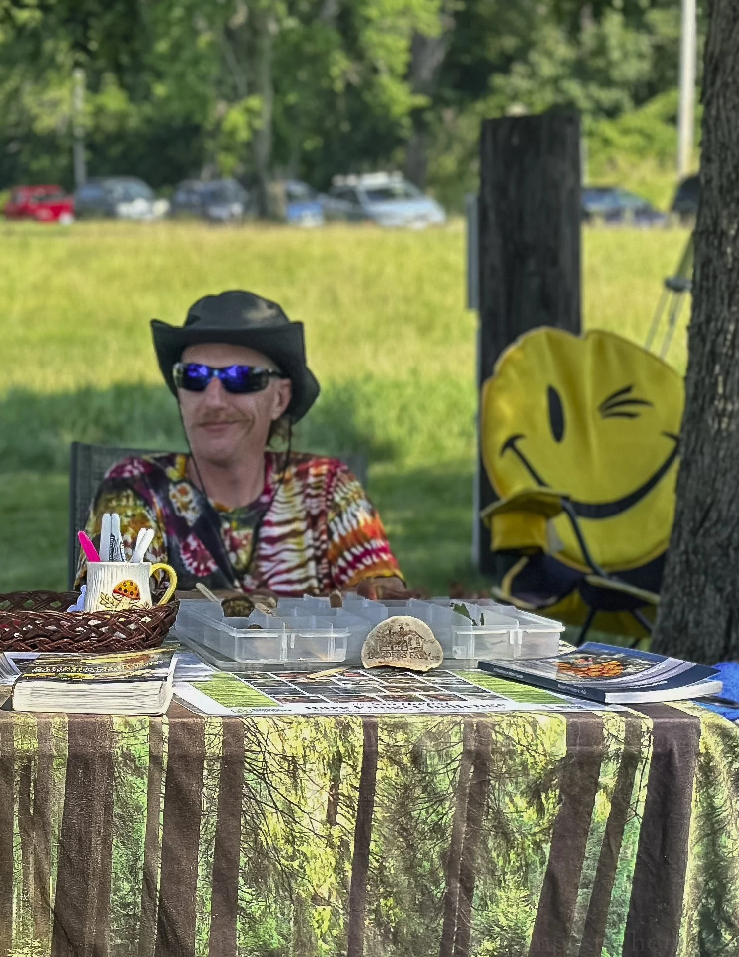 A man sitting at a table outdoors, wearing a black cowboy hat and sunglasses, with a colorful tie-dye shirt. There is a yellow backpack with a smiley face winking and sticking out its tongue to the right of the man. The table has a basket with pens and a cup, a rock, and folded papers or booklets. The background shows a grassy area with trees and a few parked cars.