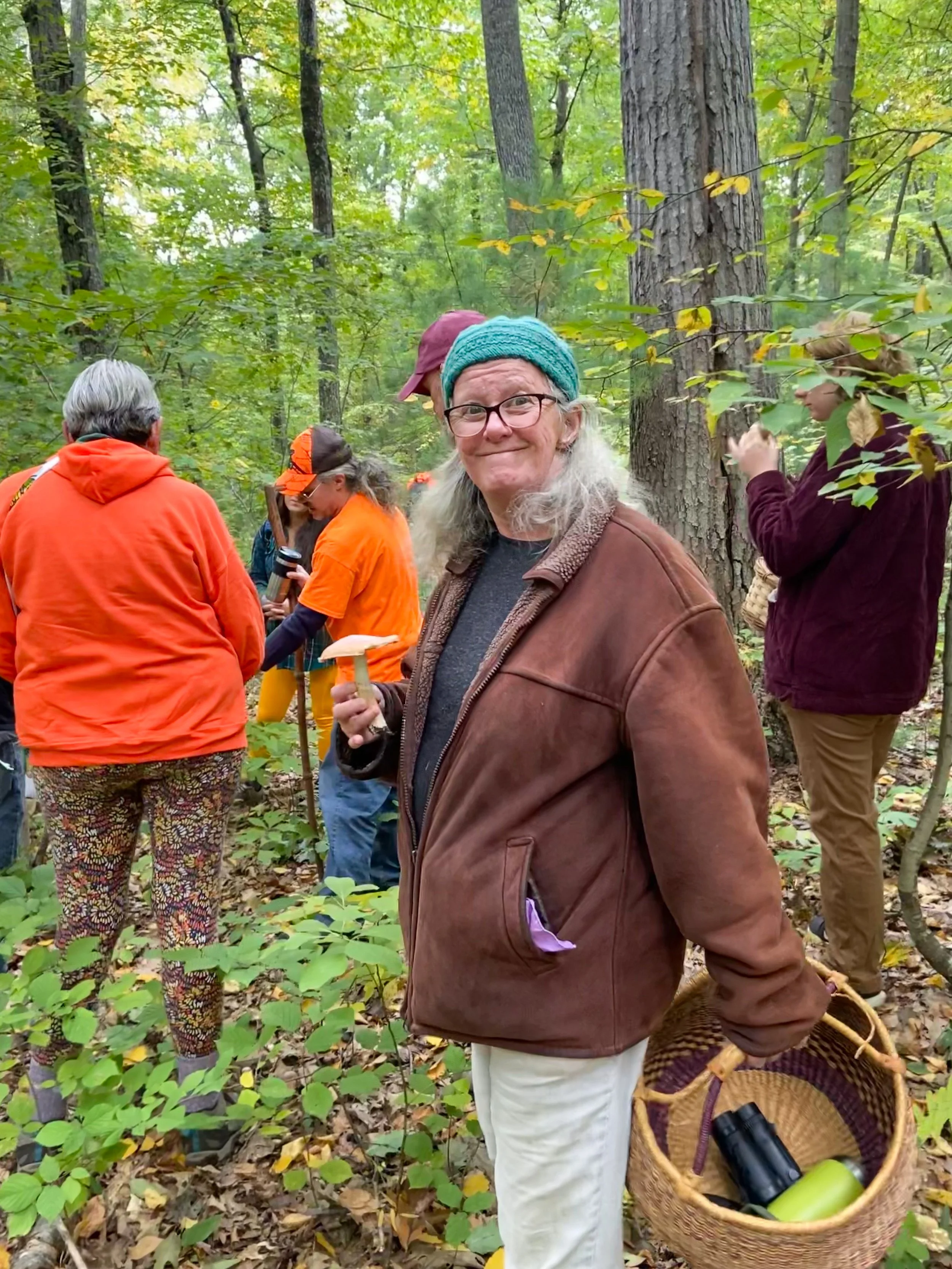 A woman with gray hair, glasses, a teal knit cap, and a brown jacket holding a mushroom, standing in a forest with other people. Some are wearing bright orange clothing, possibly for foraging. The woman is smiling and holding a basket with binoculars and a green water bottle inside.
