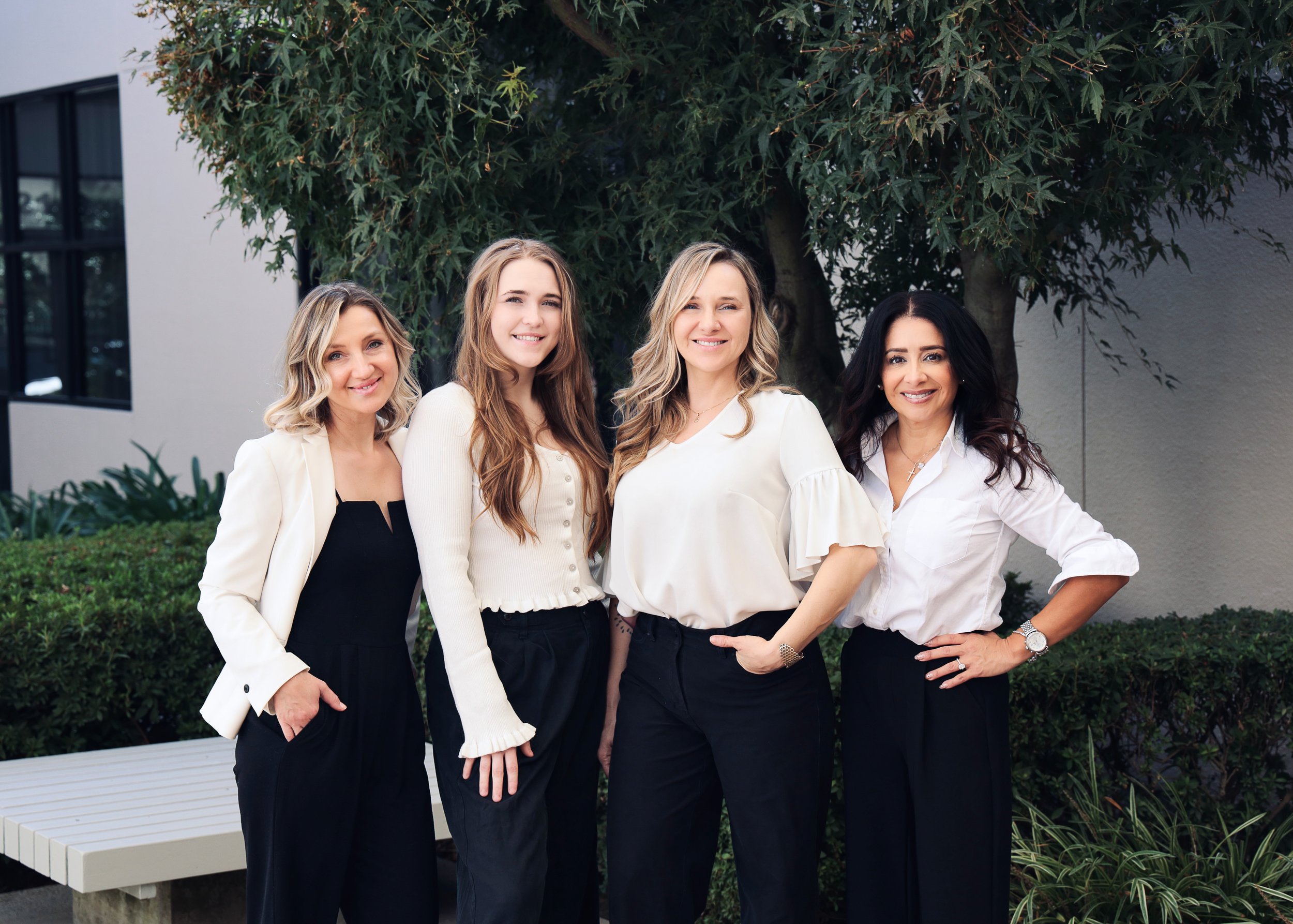 Four women standing outdoors in front of a tree and bushes, dressed in professional attire, smiling at the camera.