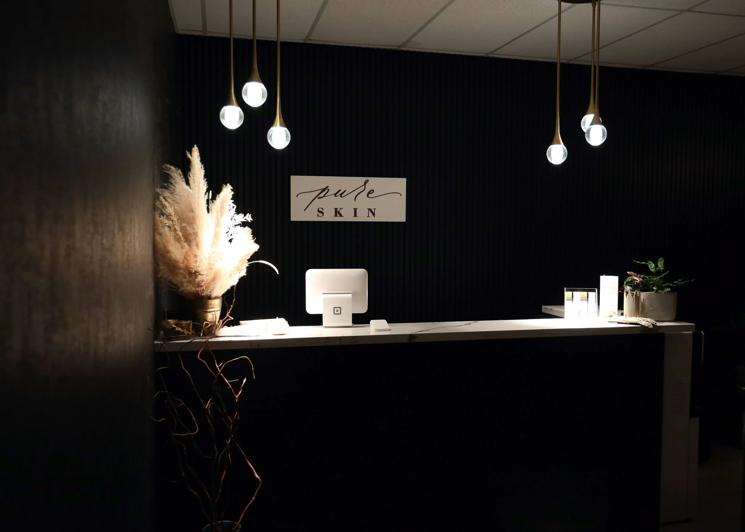 Reception desk with a computer, potted plant, and decoration, behind a dark wall with a "Pure Skin" sign, and hanging modern pendant lights.
