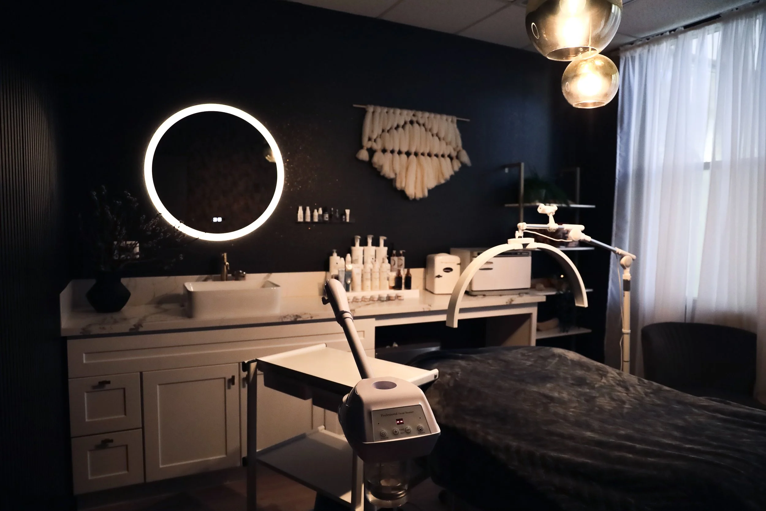 A modern aesthetics treatment room with a dark wall, illuminated round mirror, and a treatment bed covered with a dark blanket, surrounded by skincare products, equipment, and a window with white curtains.