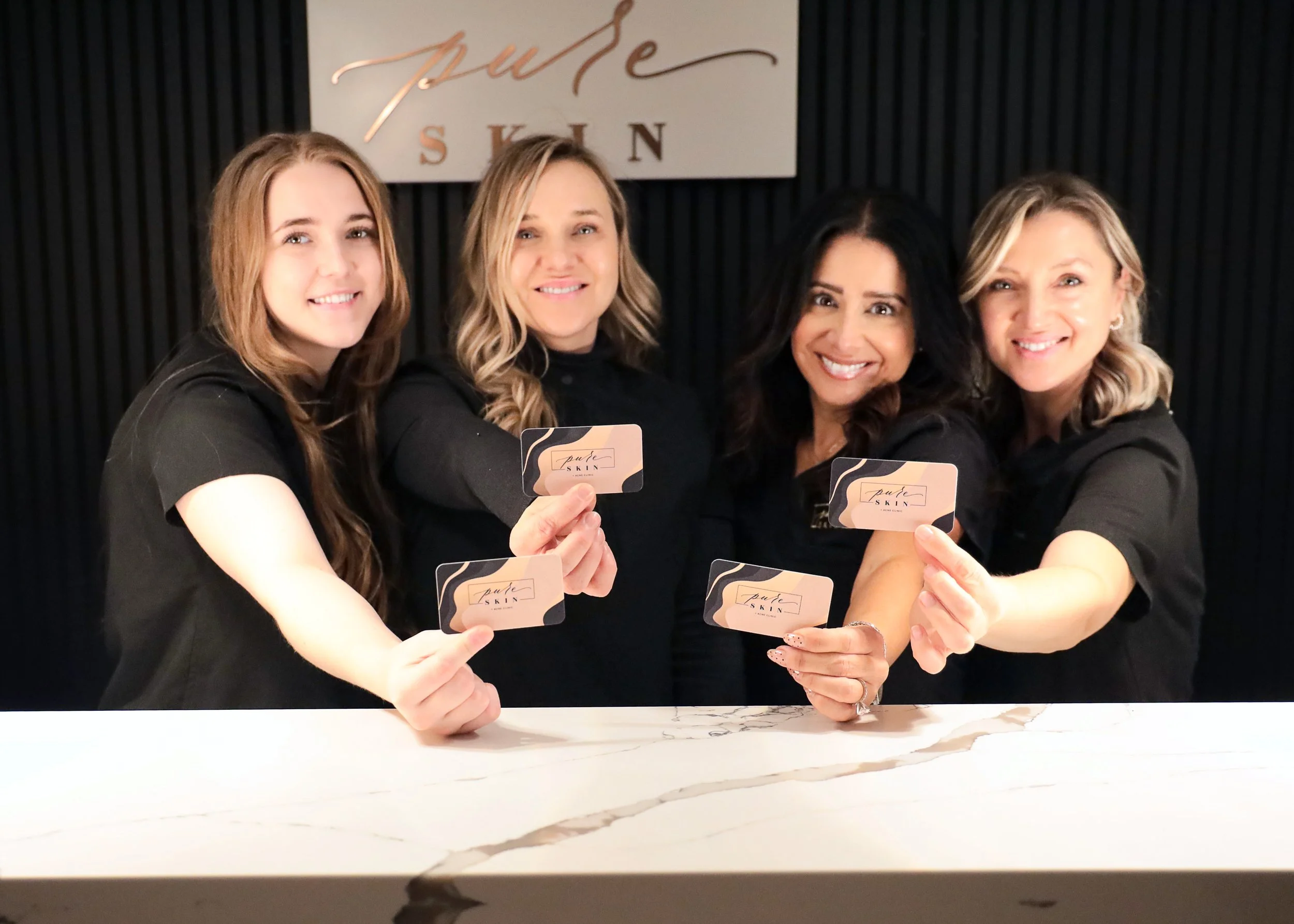 Four women holding business cards with the logo "Pure Skin" in a salon or spa setting, smiling at the camera.