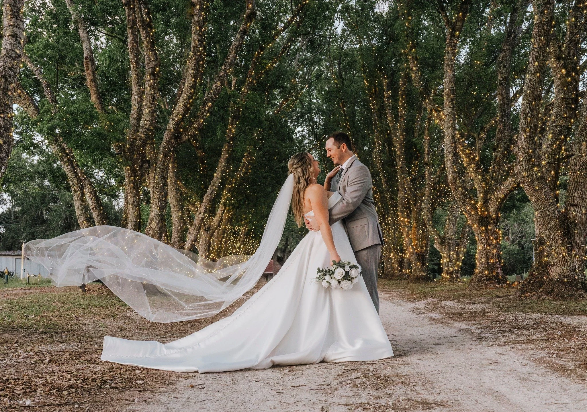 Bride and Groom hugging in the middle of a driveway at the wedding venue. Behind them are tall trees wrapped in lights. The Bride is holding her bouquet on one hand. 