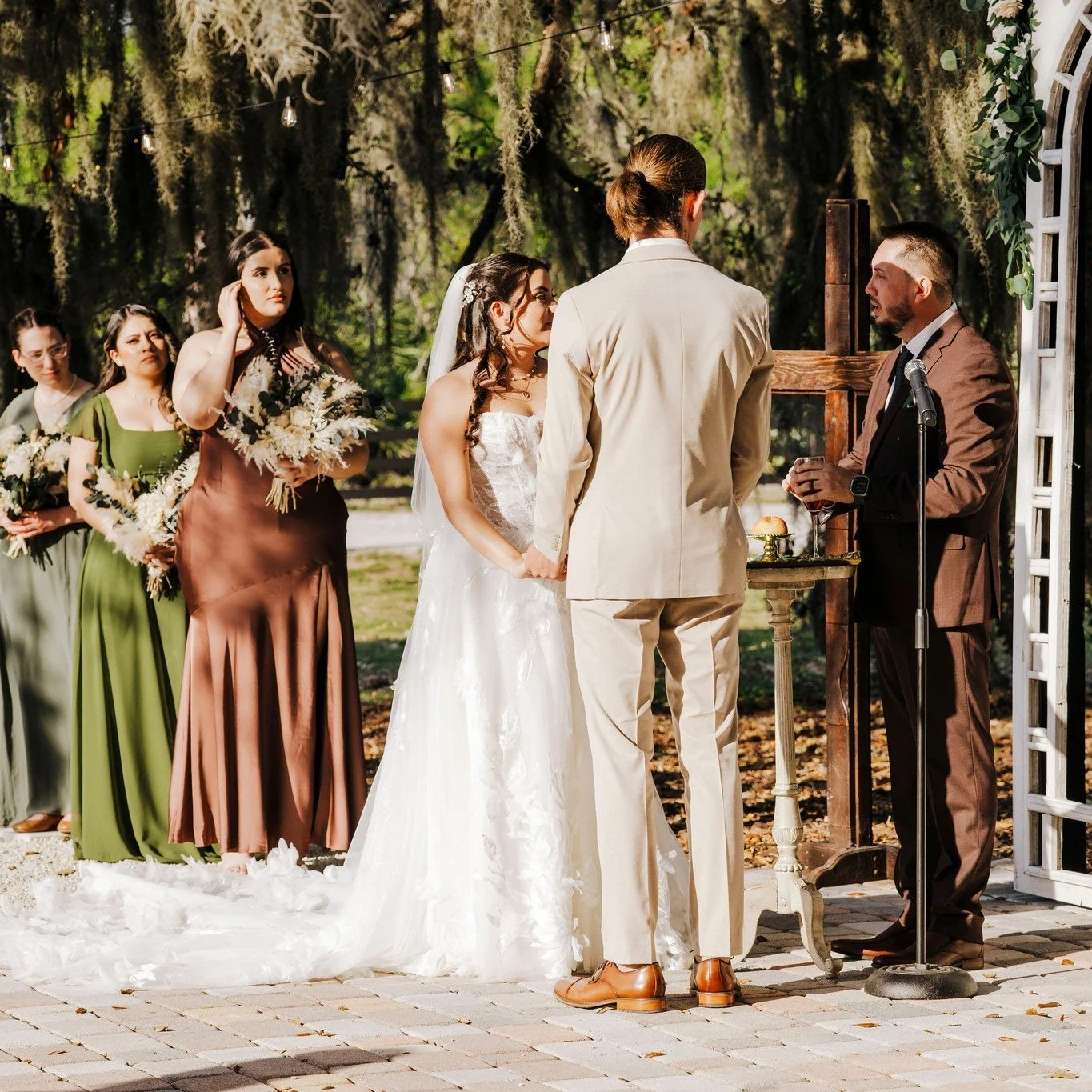 Bride and groom sharing vows during wedding ceremony. They are holding hands and are standing by the alter. Behind the bride is her bridal party. 