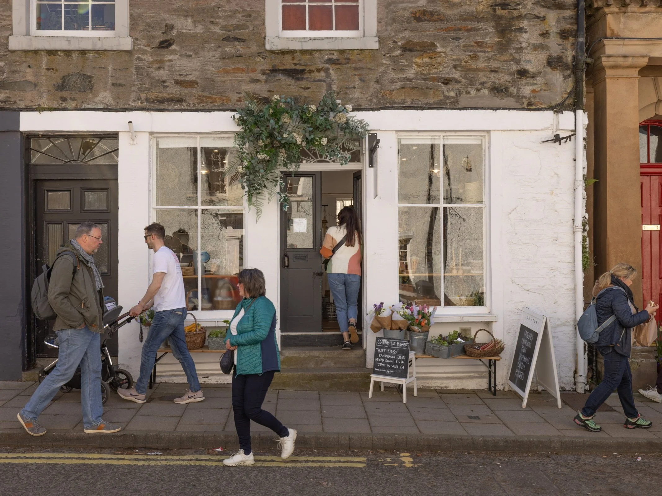 People walking past a storefront with large windows and a door, decorated with a bouquet of greenery above the entrance, on a city street.