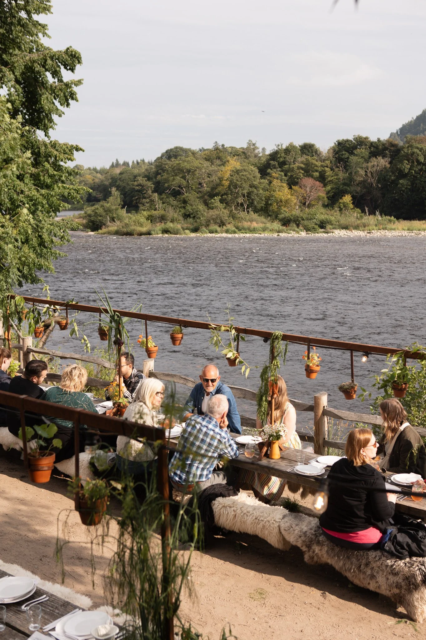 A scenic view of a small town situated along a river, with a stone bridge crossing the water, surrounded by lush green trees and hills under a clear blue sky.