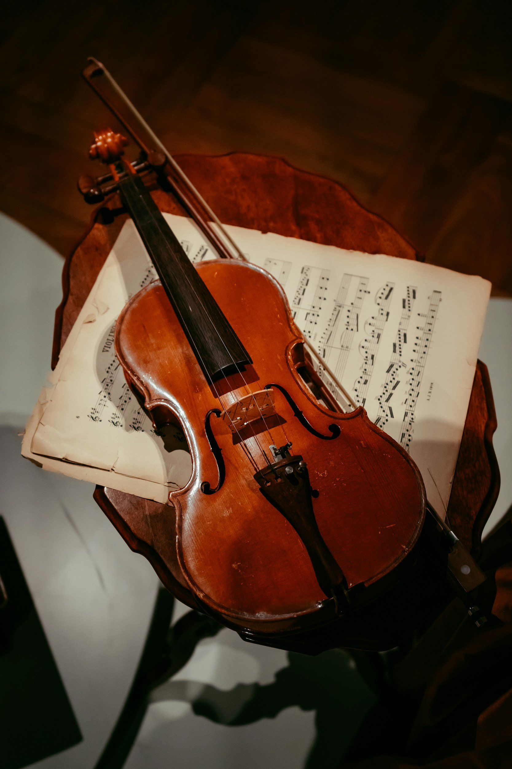 A wooden violin resting on a sheet of music on a small table, with a cello bow crosswise on top of the violin, in a dimly lit room.