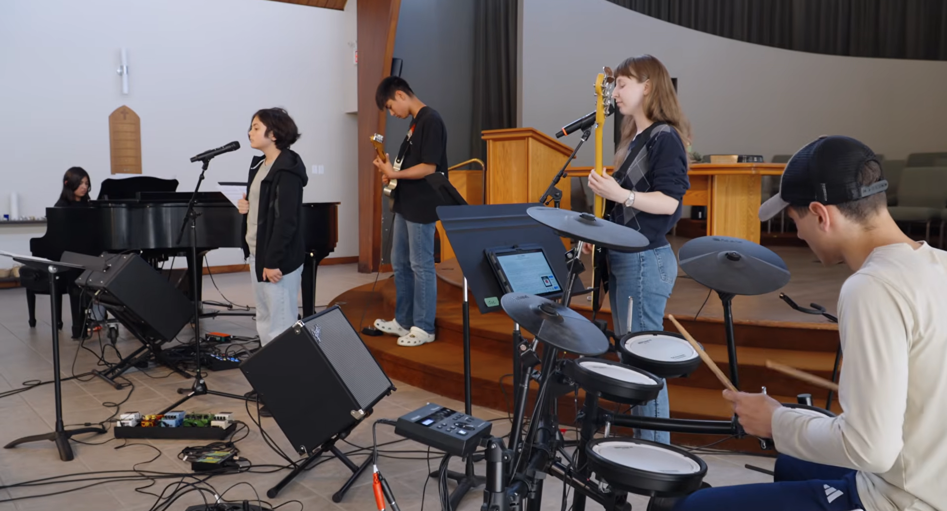 A group of young girls playing musical instruments and singing in a recording studio. One girl stands in front of a microphone, singing or speaking, while three others hold guitars and sit with music stands.