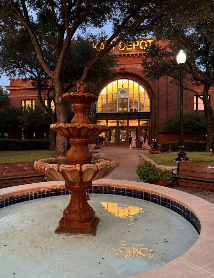 The lighted Katy Depot sign and arched window behind the front fountain at dusk in Denison, Texas