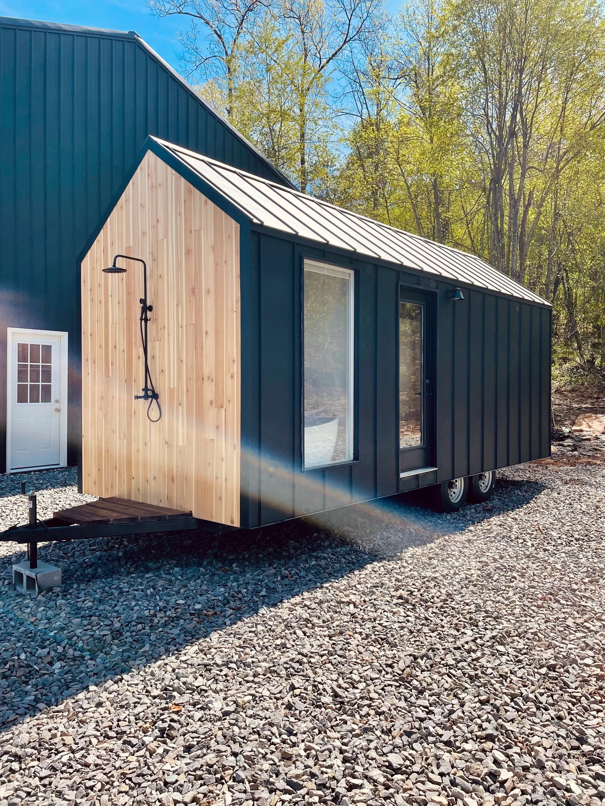 A tiny house with black metal siding and a natural wood section, with large windows and a metal roof, situated on a gravel surface surrounded by trees and blue sky.