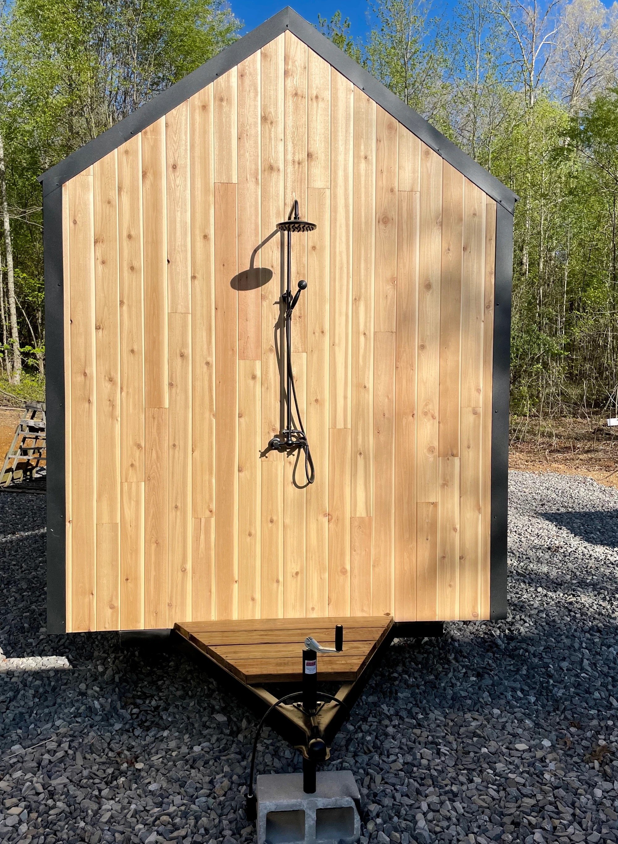 An outdoor shower with a wooden wall, black trim, and a metal shower head mounted on the wall. The shower is on a trailer with a gravel foundation, surrounded by trees and a blue sky.